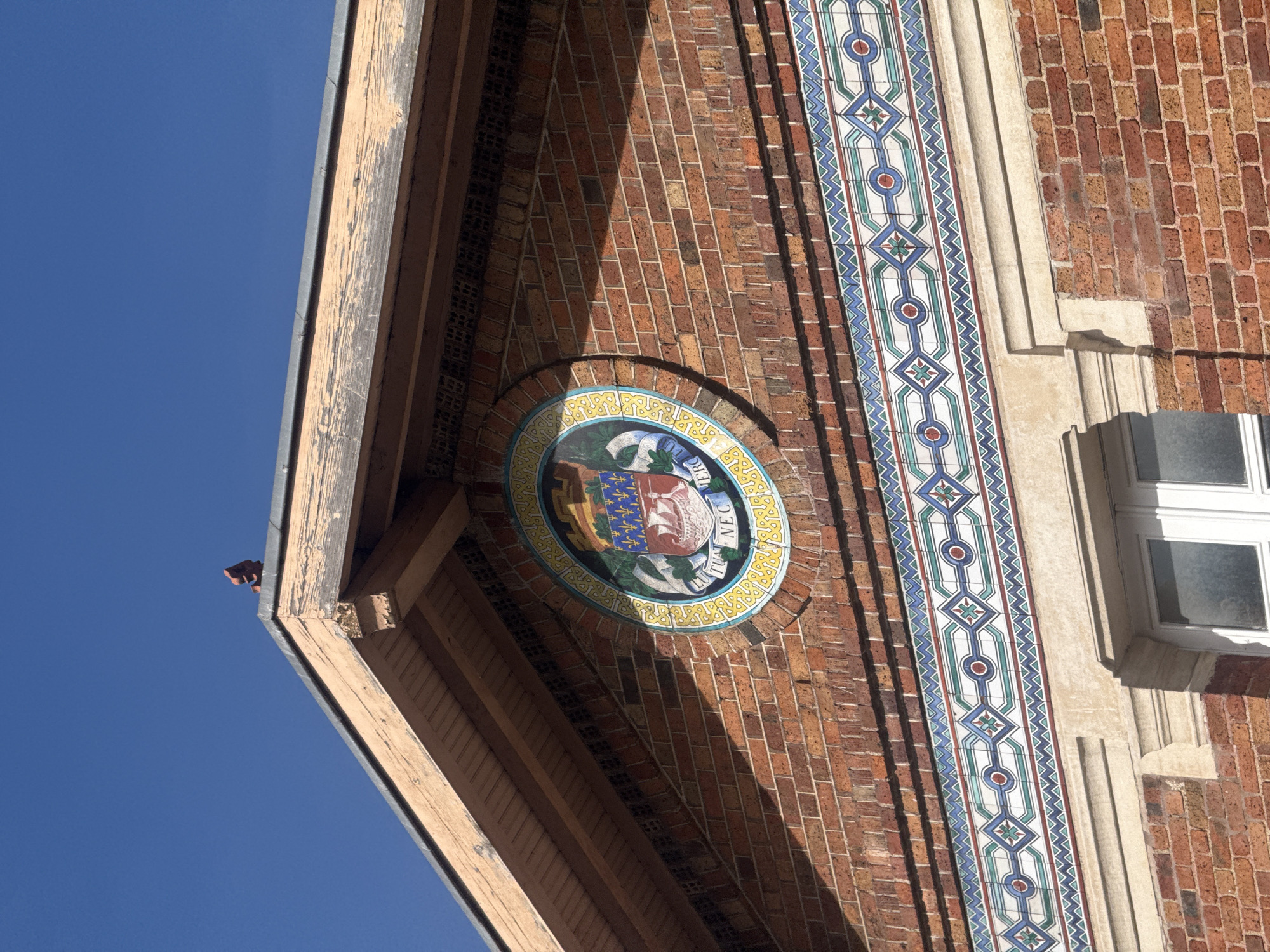 A low-angle shot depicts a section of a brick building in Paris, France, under a clear blue sky. The building features a decorative, circular emblem with a coat of arms, inscribed with "FLUCTUAT NEC MERGITUR," a Latin phrase meaning "She is tossed by the waves, but does not sink," which is the motto of Paris. This emblem is centered on a brick façade, above an ornamental tile design that runs vertically. To the right of the tile design, a partially visible white-framed window is present. The building's roofline, constructed of timber beams, angles upwards towards the left of the frame. A dark-colored object is perched at the very edge of the roof.