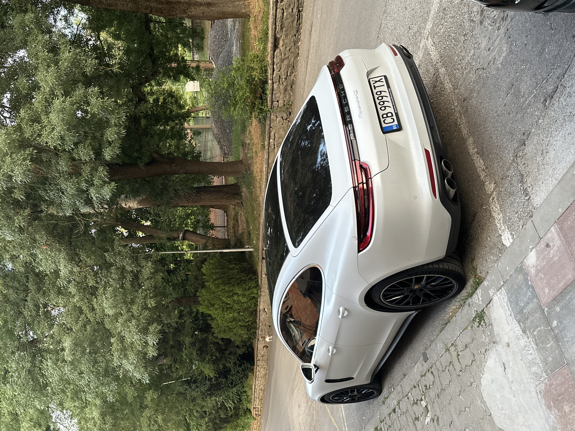 A white Porsche Panamera 4S is parked on the right side of a paved street, facing away from the camera. The car has a black roof and black rims. The license plate is visible and reads "CB 9999 TX". Behind the car, a stone retaining wall runs along the street, topped with lush green bushes. Beyond the wall, there are large trees with dense foliage, indicating a park or wooded area. In the distance, a pile of gravel and some fencing can be seen. The foreground shows a cracked concrete sidewalk with some patches of grass. A small, light-colored dog is visible on the street behind the car. The overall setting appears to be a quiet, tree-lined urban environment, likely a residential street or a park entrance in Veliko Tarnovo, Bulgaria.