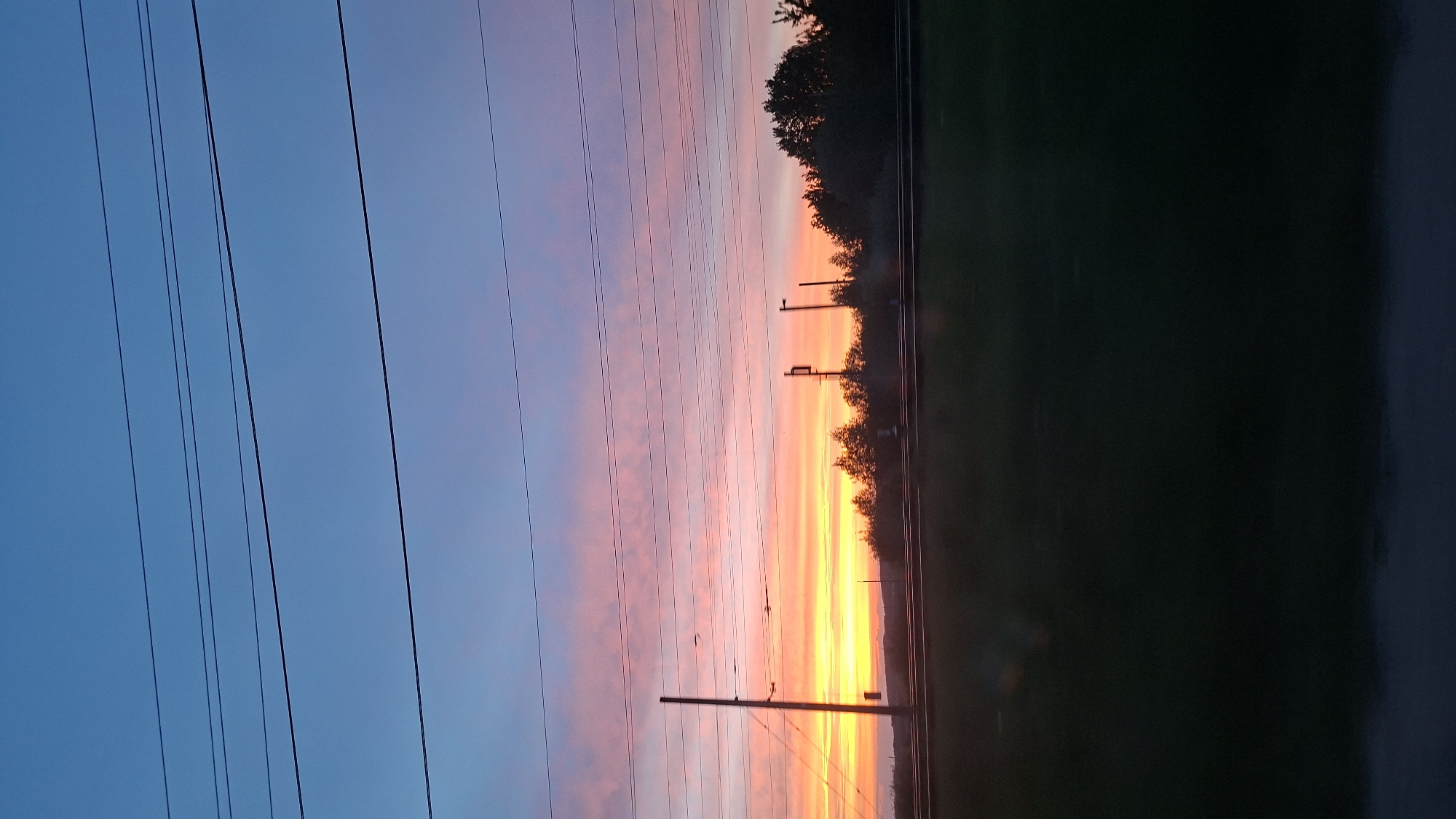 A landscape photograph taken in an unknown location shows a vibrant sunset. The sky transitions from a deep blue on the left to shades of pink and orange near the horizon. High-voltage power lines run vertically across the left side of the frame, adding a linear element to the composition. In the center, utility poles are visible. A dark green field or vegetation occupies the right side of the frame. The absence of people or specific identifiers makes it difficult to pinpoint the exact location within the city of Unknown.