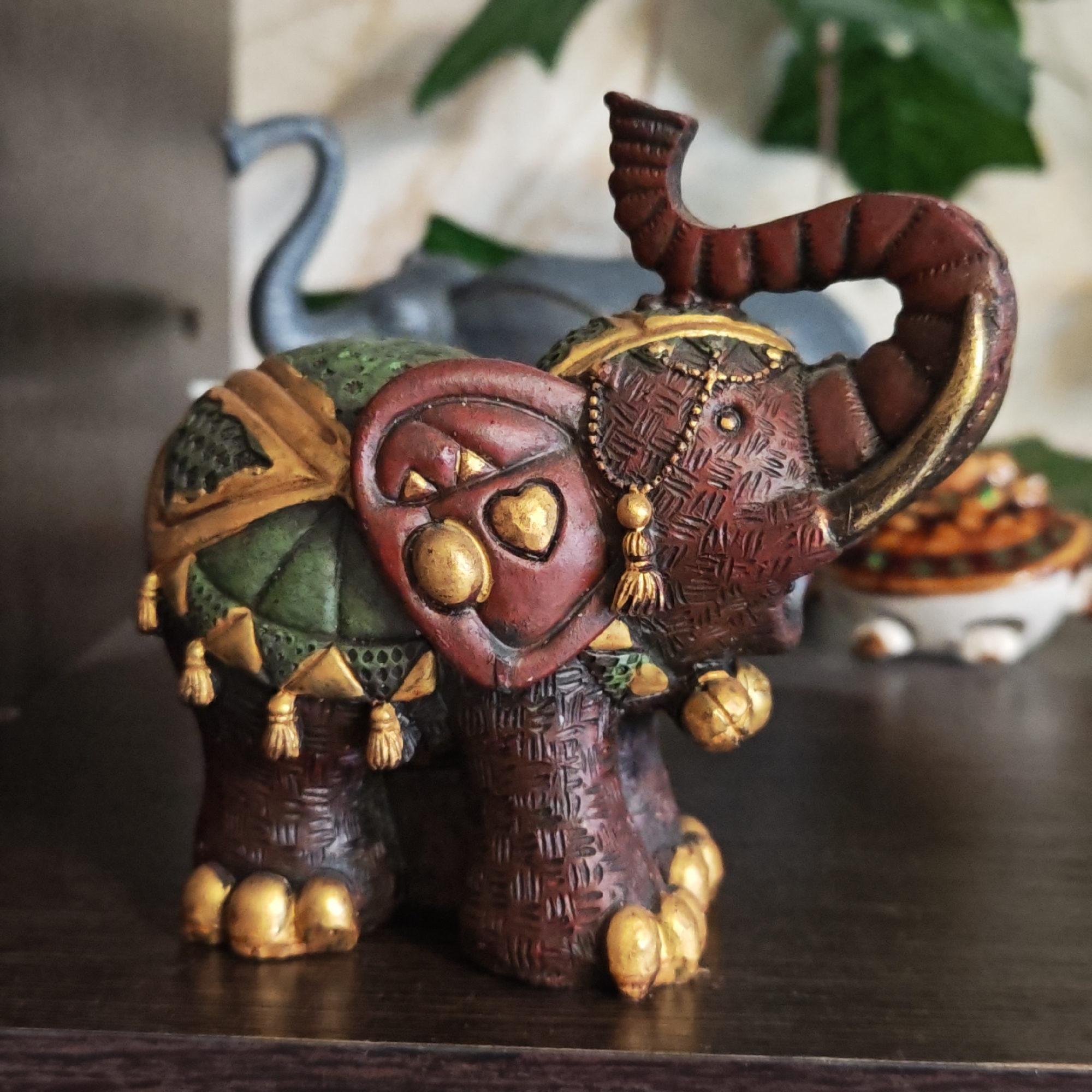 A close-up photograph shows a decorative, ceramic elephant figurine on a dark surface. The elephant has a maroon-colored body with detailed textures that mimic woven material. Gold accents highlight the feet, ear decorations, and tassels hanging from the body covering. The body covering itself is primarily green with gold edging. The elephant's trunk is raised. Another blurred, gray elephant figurine is visible in the background, along with a green plant. A small decorative container sits to the right of the elephant figurine. The environment suggests an indoor setting, possibly a shelf or display area. The image location is Novo-Nikol'skoe, Russia, but there's no indication or verification in the image itself.