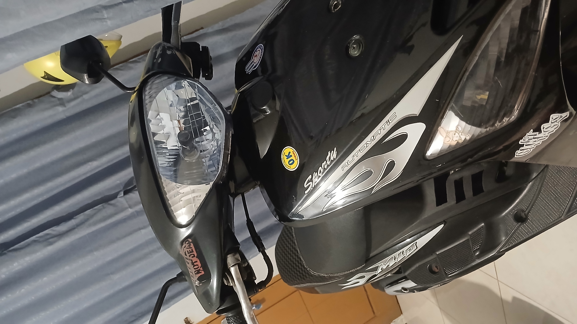 A black Yamaha Mio scooter is parked indoors, possibly in a residential setting in Sampang, Indonesia. The scooter has several stickers, including one that reads "HOT DEALS" and another circular one with "OK YAMAHA" printed on it. The scooter's headlight is clearly visible, as are its handlebars and part of the seat, which has a carbon fiber pattern. A yellow helmet is perched on a black rearview mirror on the right side of the scooter. The background consists of a grey curtain and a portion of a wooden door. The lighting suggests an indoor environment, possibly dimly lit or at night.