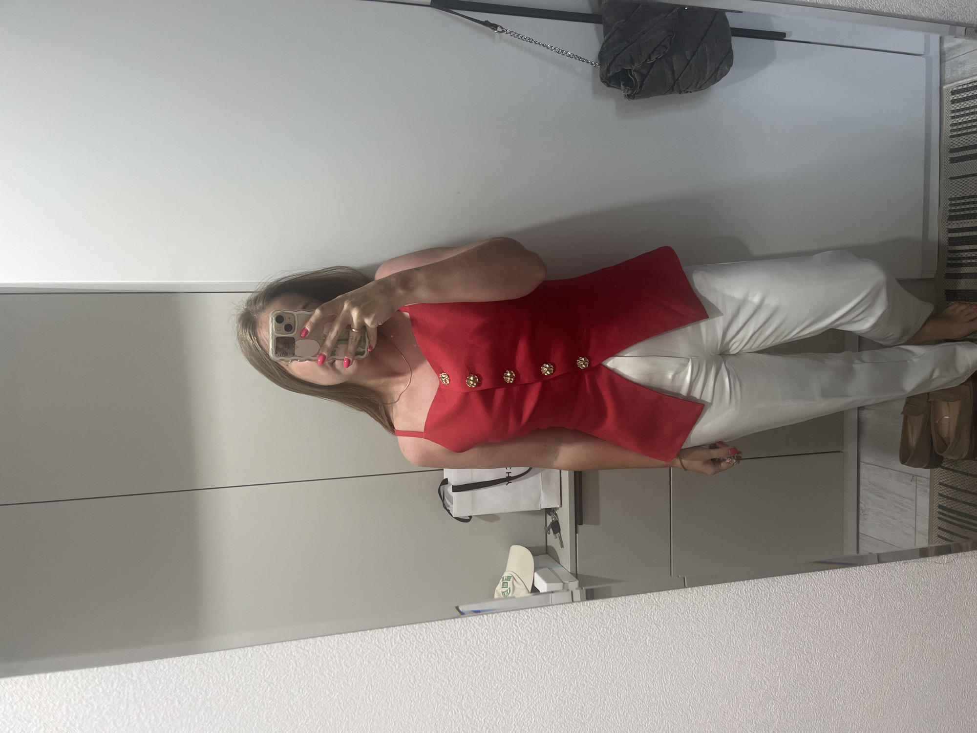 A person, viewed from the shoulders up, is taking a mirror selfie. They are wearing a red sleeveless top with gold buttons and white pants. Their right hand is holding a smartphone, obscuring their face, and their nails are painted a bright pink. The background is a white wall with a framed picture or mirror above and a closet or cabinet below. To the right of the frame, there's a patterned rug and a pair of brown shoes. A black quilted purse with a chain strap hangs on the wall. A white shopping bag with keys is on a shelf below the mirror. The setting appears to be indoors, possibly a bedroom or dressing area. The lighting is bright, suggesting daytime. No weather or specific emotional cues are evident. No visible text.