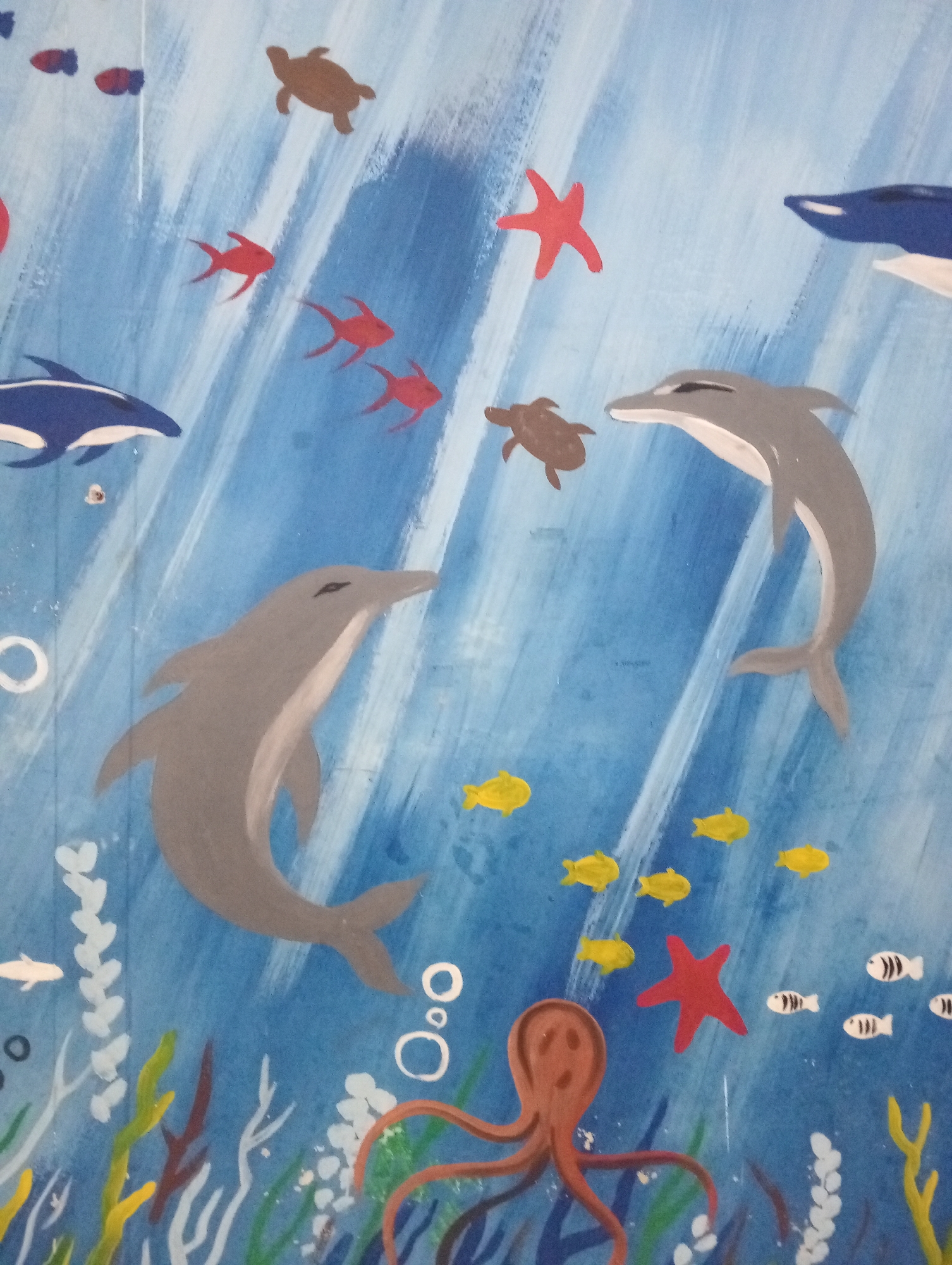 The image depicts a mural of an underwater scene, likely painted on a wall. The mural features various marine life, including two dolphins, one grey and one blue and white, swimming amidst red and yellow fish, a brown sea turtle, and a red starfish. An orange octopus is visible at the bottom, with its tentacles spread out. Bubbles are depicted as white circles of varying sizes. The background is a blue gradient with white streaks suggesting light rays or water movement. The style of the painting is simplistic and illustrative. There are no people visible in the image. The mural appears to be located in Pringsewu, Indonesia, possibly on a building or a public space, given the context of murals often being found in such locations.