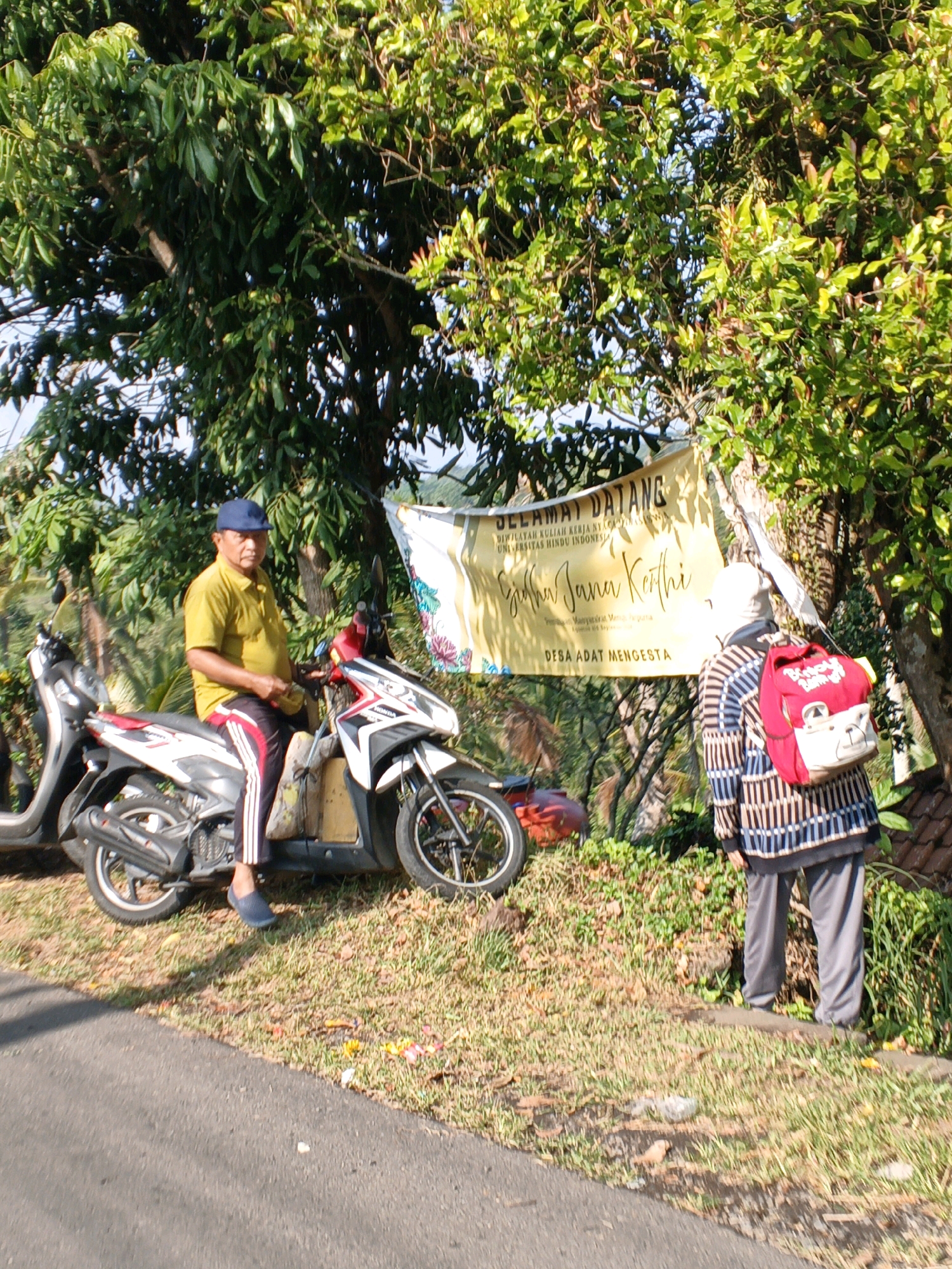 In the city of Penebel, Indonesia, a man wearing a yellow t-shirt and a blue cap sits on a motorcycle parked on the side of a road. He is looking forward, and his hands are resting on the handlebars. To his right, a banner hangs on a tree, with Indonesian text that reads "SELAMAT DATANG" (Welcome) and "DESA ADAT MENGGSTA" (Mengesta Traditional Village). Another person, wearing a striped long-sleeved shirt and grey pants, stands facing away from the camera, looking at the banner. They are carrying a red backpack with a bear design. The scene is surrounded by lush green trees and vegetation, and the sunlight suggests it is a clear day.