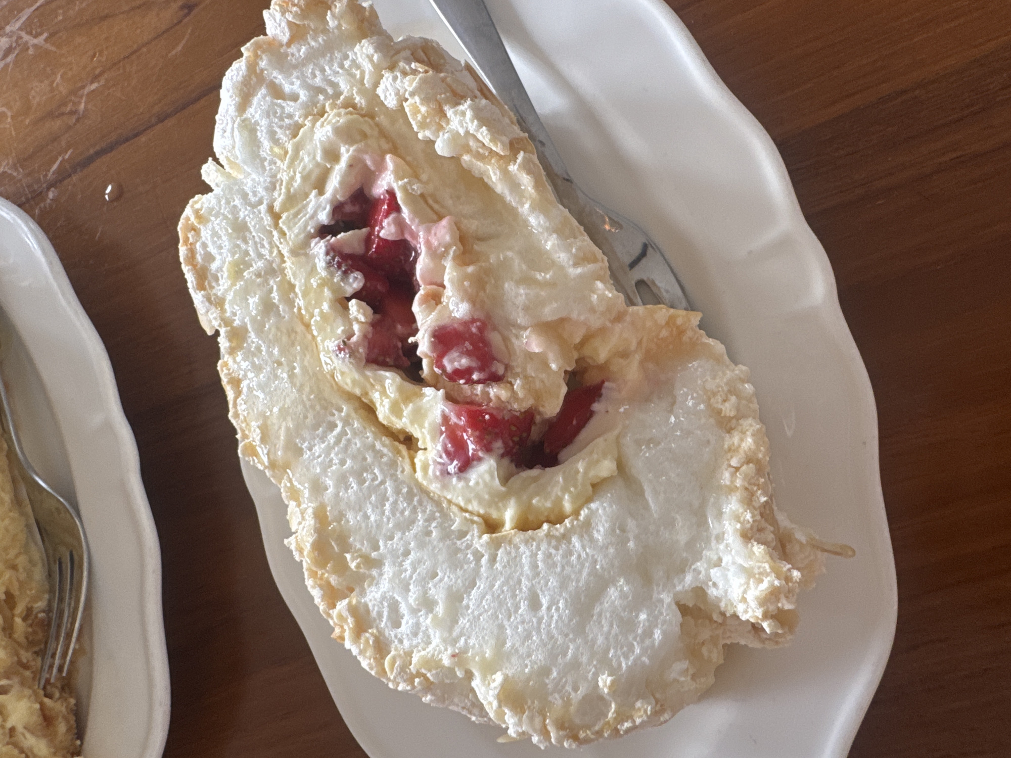 A slice of meringue roulade with fruit filling is presented on a white plate. The roulade is made of a light, airy meringue base, rolled around a creamy filling and chunks of red fruit, possibly strawberries or raspberries. A fork is visible on the right side of the plate, partially submerged in the dessert. The plate sits on a wooden table, suggesting an indoor setting, possibly a cafe or restaurant. The lighting is soft, highlighting the textures of the meringue and the fruit. The image is taken from a slightly elevated angle, focusing on the dessert. The location of Kuta Selatan, Indonesia is not discernible from the image itself.