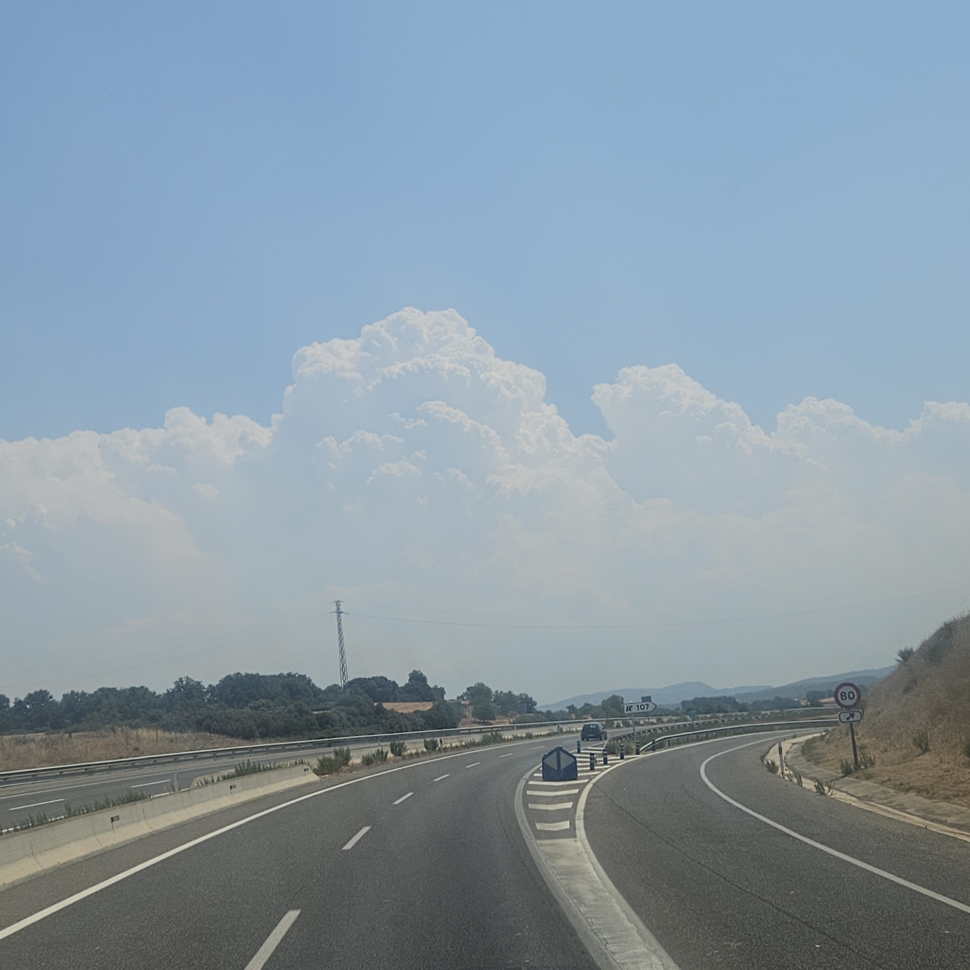 The image captures a view from a moving vehicle on a highway, likely in or near Calaf, Spain. The sky is a clear blue with large, puffy white clouds dominating the upper portion of the frame. In the middle ground, a landscape of green trees and vegetation lines the side of the road, with a tall power pylon visible in the distance. The highway itself curves to the right, with clear white lane markings and a concrete barrier separating the lanes. A speed limit sign indicating 80 is visible on the right side of the road, along with a road sign pointing to "R 107." A few vehicles are present on the highway, including a dark-colored car in the distance and a glimpse of another vehicle in the foreground. The overall environment suggests a sunny day on a rural or semi-rural highway.