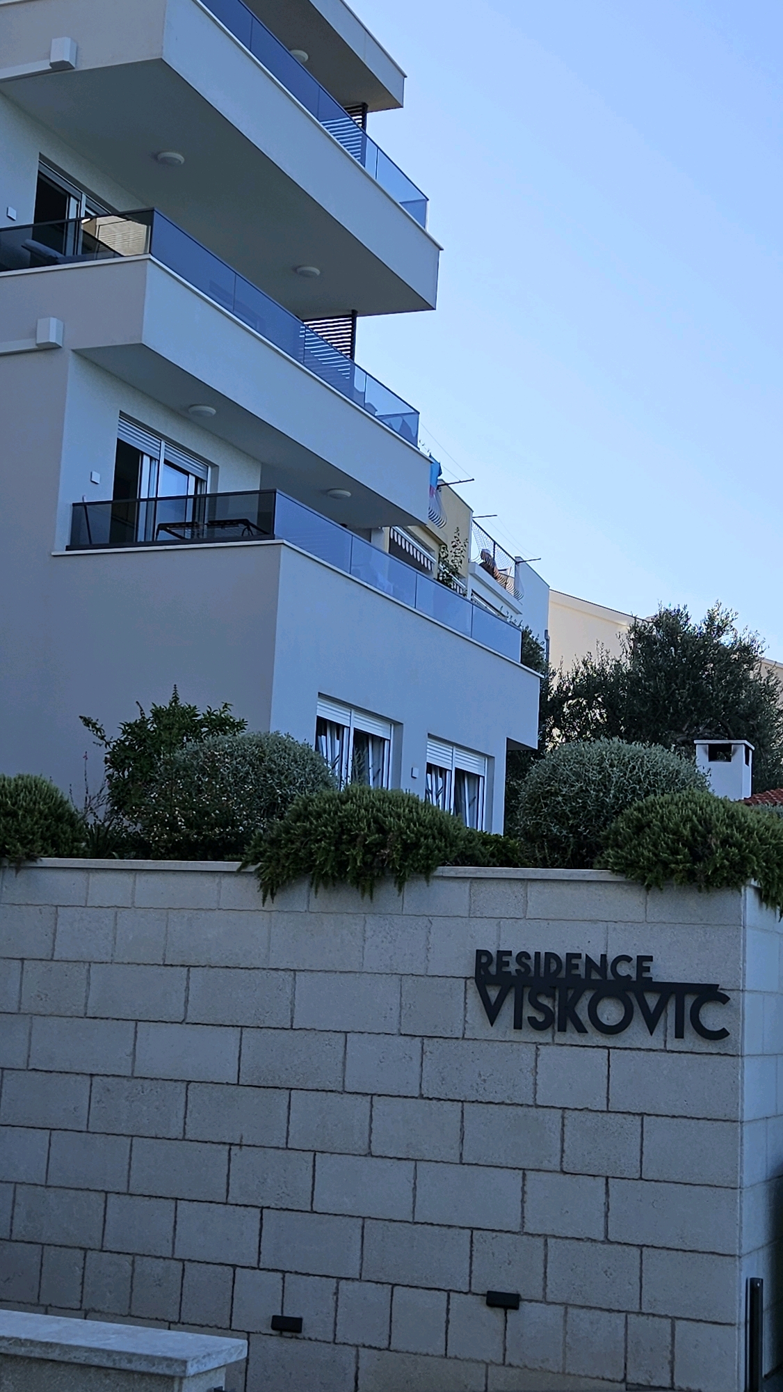 The image shows the exterior of a modern apartment building in Općina Tučepi, Croatia. The building has multiple balconies with glass railings and a light-colored facade. In front of the building, there is a low, light-colored stone wall with the words "RESIDENCE VISKOVIC" inscribed in black metal letters. Several green bushes are planted in front of the wall, and a small chimney is visible on the right side of the image. The sky is clear and blue. There are no people clearly visible or identifiable in the foreground, although figures can be faintly seen on a balcony in the upper right portion of the building.