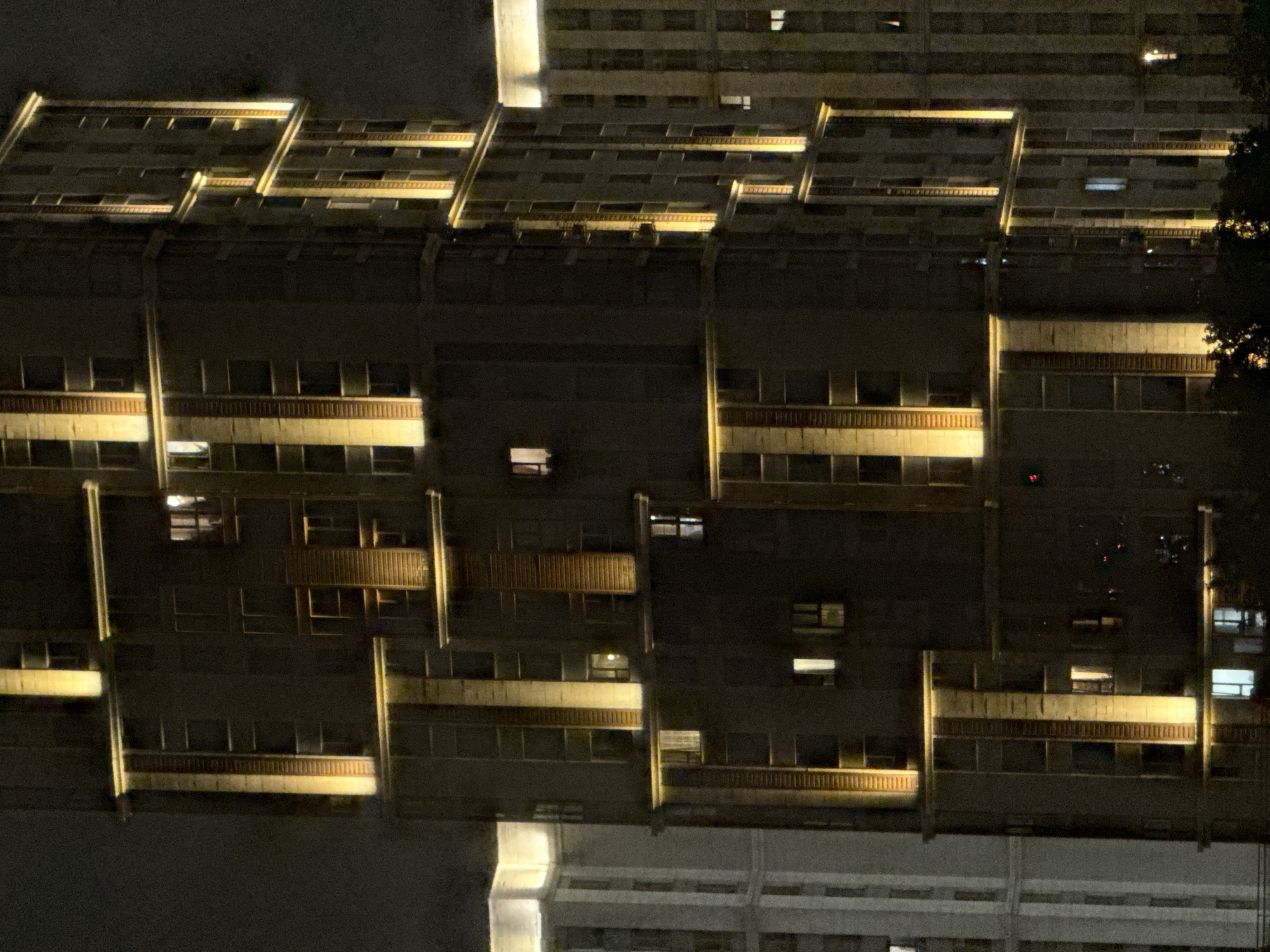 The image shows a nighttime view of modern apartment buildings in the city of Khimki, Russia. The buildings are illuminated by external lights, highlighting their architectural features. The main focus is on a tall, dark-colored building with vertical and horizontal bands of warm yellow light. This building has numerous windows, some of which are lit from within, suggesting occupancy. To the left of this building is another lighter-colored building, also featuring exterior lighting. The sky is dark and appears overcast, typical of nighttime in a city. Trees are visible in the foreground, silhouetted against the lights of the buildings. The scene depicts an urban environment at night, characterized by residential architecture and artificial illumination.