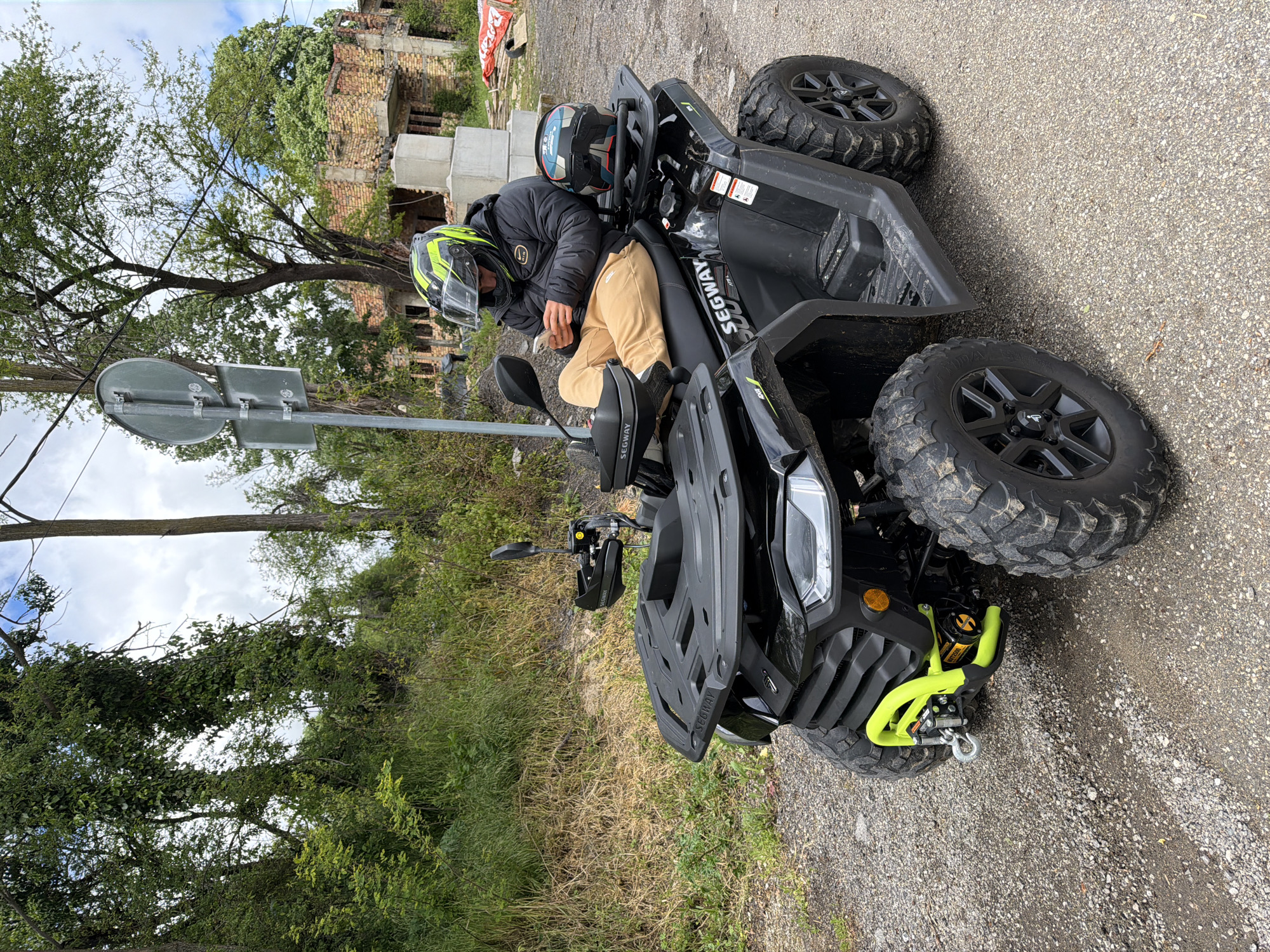 A person wearing a black jacket, tan pants, and a yellow and black helmet is seated on a black Segway ATV. Another helmet rests on the ATV's seat. The ATV is parked on a gravel road next to a patch of dry grass and a road sign. In the background, there are trees and a building that appears to be under construction or partially damaged. The location is in the city of Брусовeте, Bulgaria.