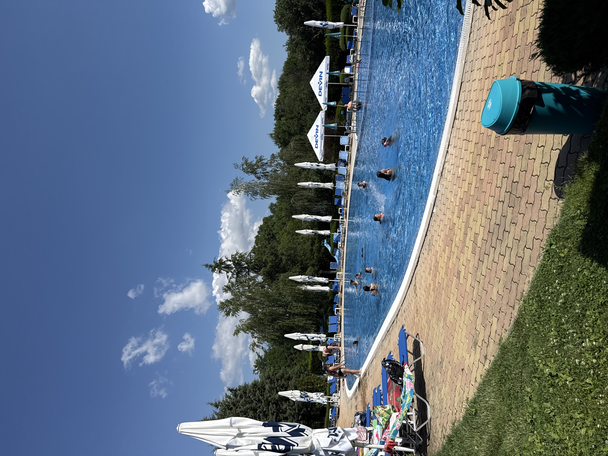 This is a horizontal, eye-level shot of an outdoor swimming pool area in Yagoda, Bulgaria. The bright blue swimming pool takes up a significant portion of the frame, with several people swimming or wading in it. The pool is surrounded by a tiled walkway in shades of yellow and red, and beyond that, a strip of green grass. A line of lounge chairs and white umbrellas with blue accents are positioned along the edge of the pool. Lush green trees and foliage form a backdrop behind the pool area. The sky is a clear, vibrant blue with scattered white clouds. The scene appears to be bathed in bright sunlight, suggesting it is a warm, sunny day, likely midday. The overall atmosphere is one of leisure and relaxation.
Visible text includes the word "DEVIN" repeated on several of the umbrellas. A large, teal-colored trash can with a black liner is also visible near the edge of the pool. The presence of lounge chairs, umbrellas, and people enjoying the water indicates a recreational setting, likely a public or resort pool.