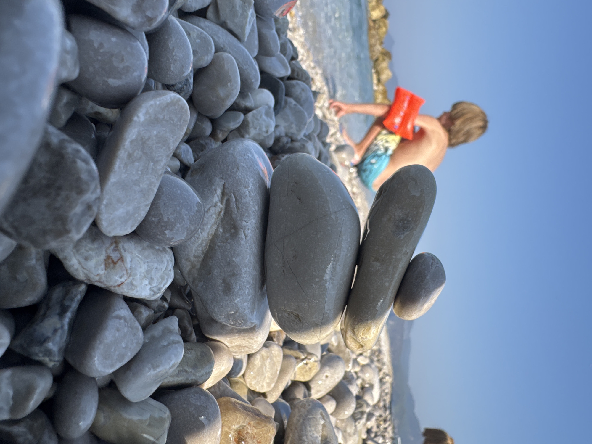 A child wearing a bright orange life vest and patterned swim trunks is playing at the water's edge on a rocky beach. The child is seen from behind, with their head slightly turned towards the viewer, and their hands are positioned to form a heart shape. The setting is a beach with numerous smooth, gray pebbles in the foreground, some of which are wet and glistening. In the background, the calm, blue water of the sea meets the shore, and beyond that, distant hills are visible under a clear, bright blue sky. The sun appears to be low in the sky, casting warm light and creating soft shadows, suggesting it is either early morning or late afternoon. The overall mood of the image is peaceful and joyful, capturing a moment of childhood play in a beautiful natural setting. There is no visible text in the image.