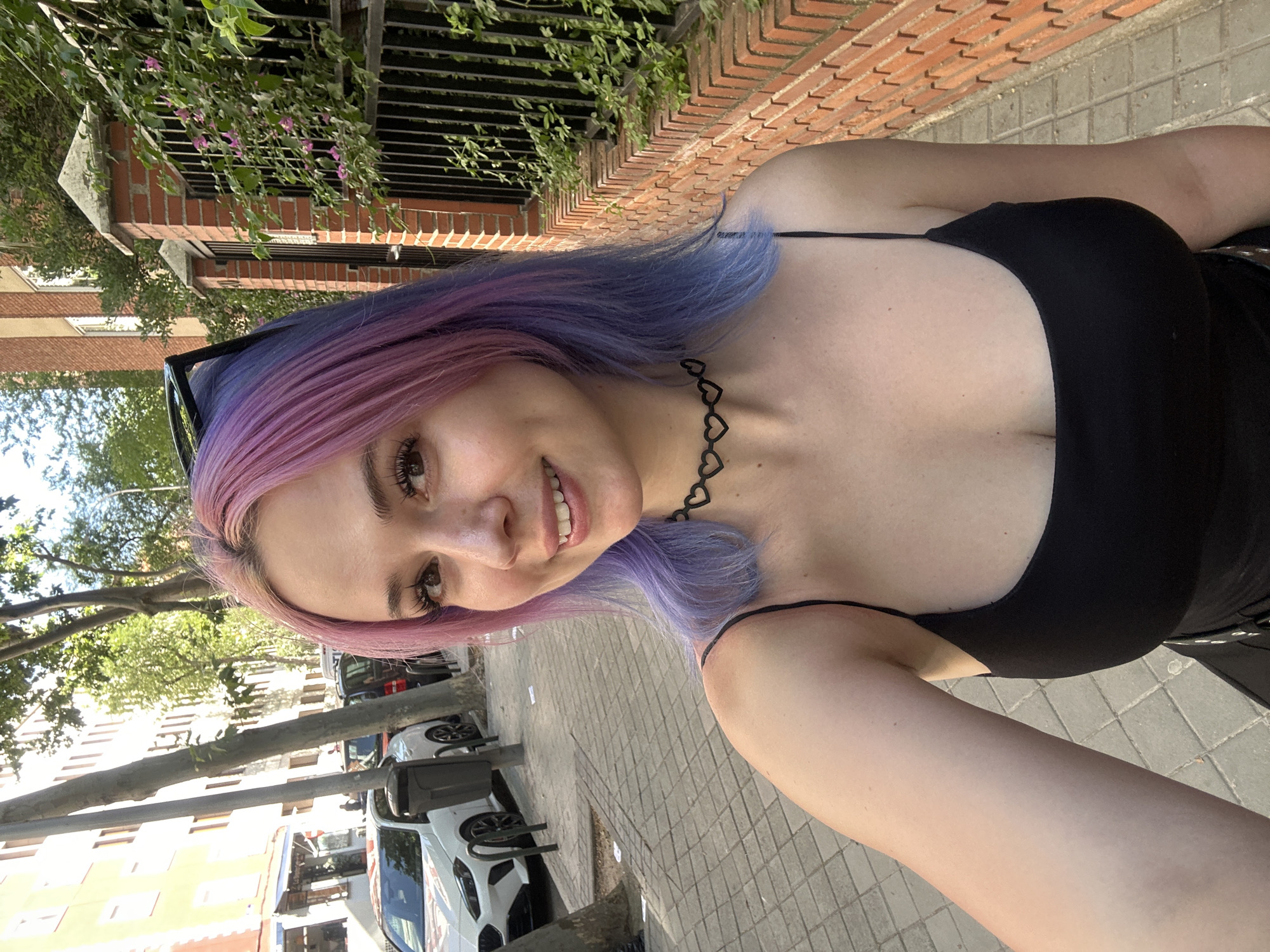 A young woman with pink and blue hair is standing on a sidewalk and taking a selfie. She is wearing a black top and a heart-shaped choker necklace. In the background, there is a brick wall, trees, and buildings, suggesting an urban environment. White cars can be seen parked on the side of the street. Based on the architectural style and the streetscape, it is possible that this photo was taken in Madrid, Spain.
