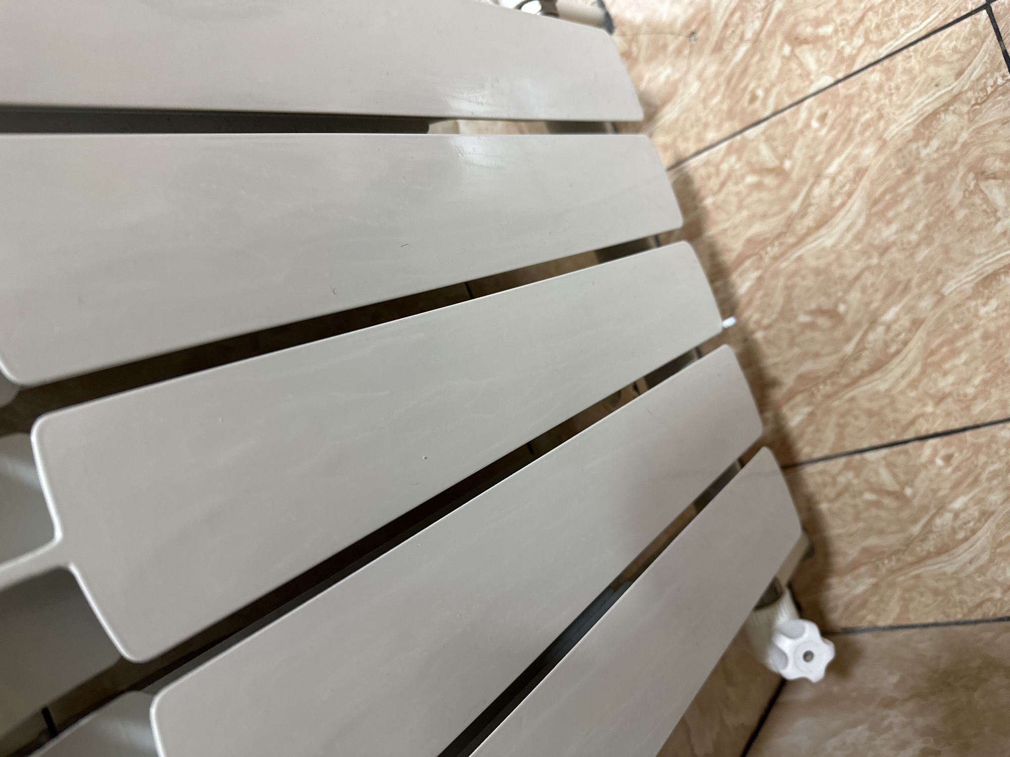 The image shows a close-up of a white radiator with vertical panels, set against a tiled floor with a marble-like pattern in earthy tones. A white valve is visible at the bottom left of the radiator. The lighting suggests an indoor environment. There is no indication of the city of Fergana City, Uzbekistan, or any specific location. The scene is static and devoid of human presence or activity.