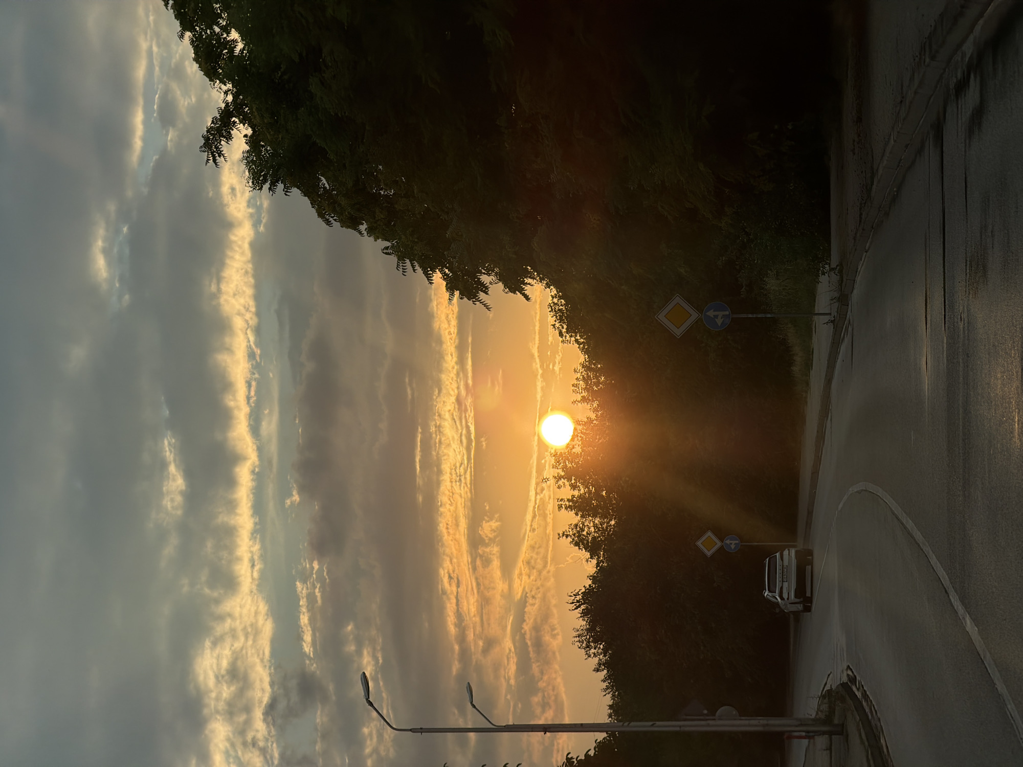 The image shows a road in Lovech, Bulgaria, at sunset. The sun is low on the horizon, casting long shadows and creating a warm, orange glow. The sky is filled with dramatic clouds, tinged with yellow and orange from the setting sun. On the left side of the road, there are two streetlights. A white car is driving away from the camera, heading down the winding road. There are trees lining the right side of the road, their leaves dark against the bright sky. Road signs, including a diamond-shaped yield sign and a blue circular sign with an arrow, are visible near the roadside. The road surface appears wet, suggesting recent rain or dew. The overall atmosphere is serene and picturesque.