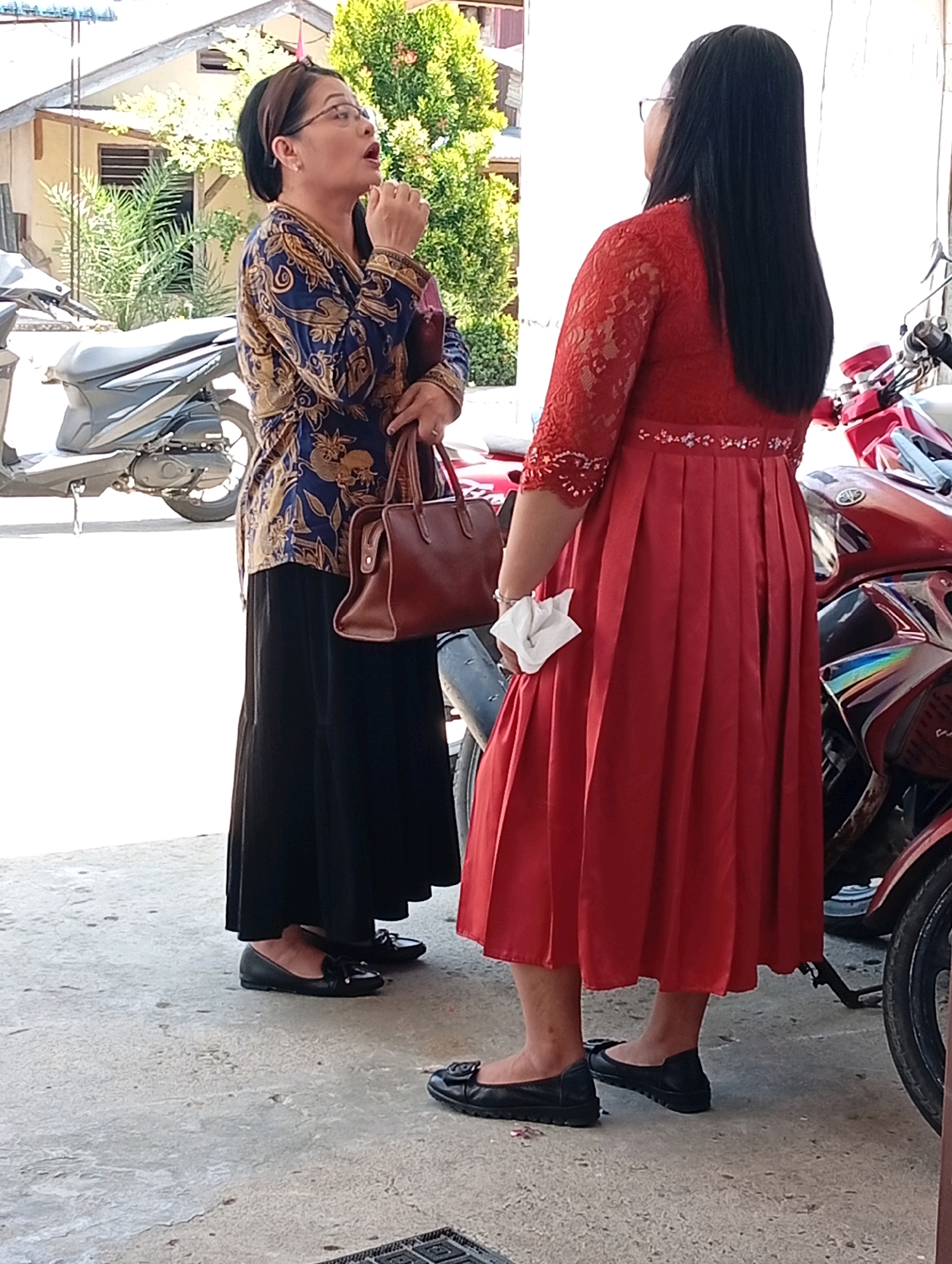 Two women are standing on a paved area in front of a building with a tiled roof. The woman on the left is wearing a blue and gold patterned top and a black skirt. She has glasses and is holding a brown handbag. The woman on the right is wearing a red dress with lace detailing on the sleeves and a beaded belt. She is holding a white tissue. Two motorcycles are visible in the background, one grey and one red. The environment appears to be outdoors, with trees and foliage visible behind the building. The scene is well-lit, suggesting it is daytime. The architecture and vegetation suggest a location in Indonesia, consistent with the city of Gunungsitoli.
