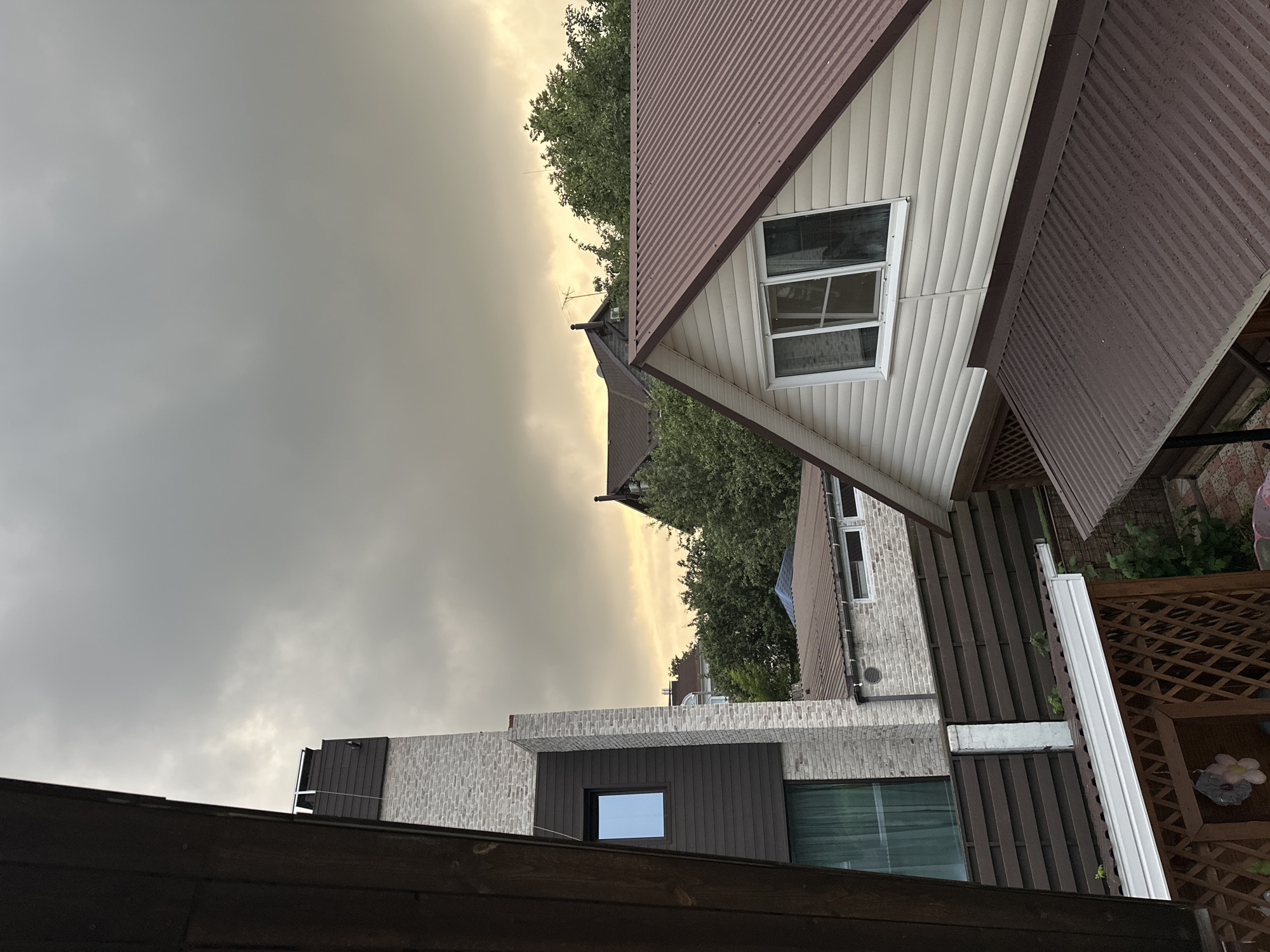 The image depicts a residential area with several houses and structures under a cloudy sky. In the foreground, a dark wooden fence or balcony railing is partially visible on the left. To the right of this, a tall, modern house features a brick lower section and a dark grey siding upper section with a small, rectangular window. Adjacent to it, a horizontal brown wooden fence runs across the mid-ground. Further back, a white house with a beige siding and a reddish-brown corrugated metal roof has a prominent gable end with a window. Behind this, a darker roof of a similar material is visible. Green trees are scattered throughout the scene, suggesting a suburban or semi-rural setting. The sky is overcast with hints of yellowish light near the horizon, indicating either dawn, dusk, or heavy cloud cover. The presence of multiple houses, fences, and varying architectural styles suggests a developed residential neighborhood. No people are visible in the image. The image appears to be a candid photograph taken from an elevated viewpoint, possibly a window or balcony. The location is identified as Nizhny Novgorod, Russia.