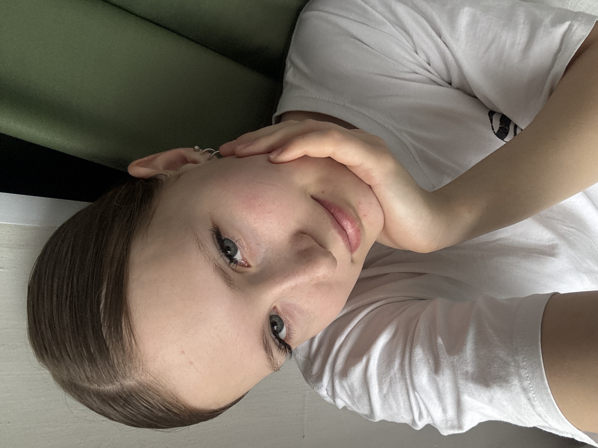 A young woman is lying down, resting her cheek on her hand. She has fair skin, dark hair slicked back, and light-colored eyes. She is wearing a white T-shirt. The background includes a green surface, possibly a sofa or wall. The location is unspecified but has been linked to Chaikovsky, Russia.