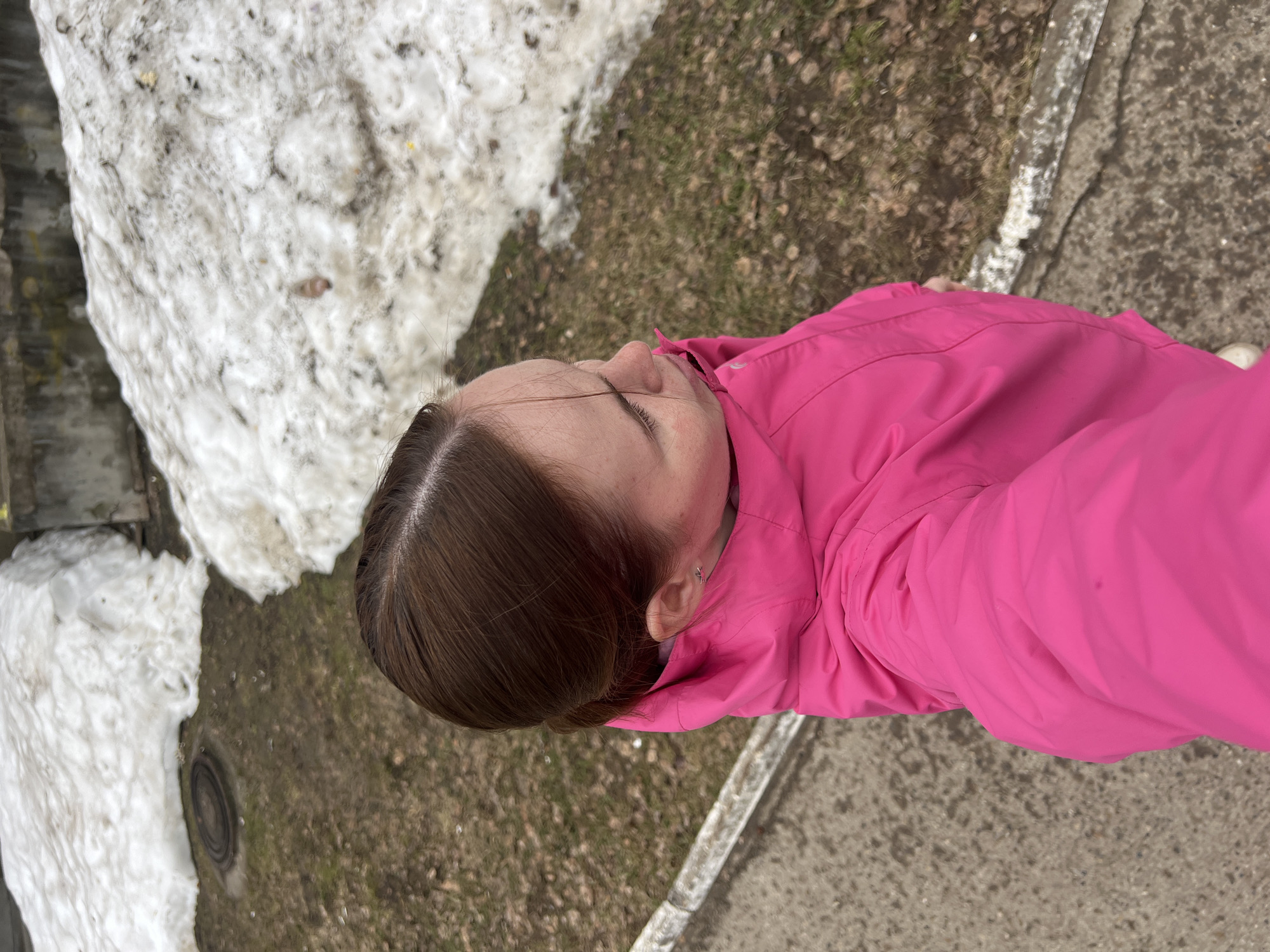 The image shows an overhead view of a person wearing a bright pink jacket. The person's head is visible, with brown hair. The person is standing outdoors.
To the left of the person, there is a patch of melting snow. Beyond the snow, there's a dark, possibly metal object, set into the ground.
To the right of the person, there's a patch of ground covered with brown foliage, possibly dead leaves or grass. A concrete sidewalk or curb is also visible.
Based on visual analysis and the provided information, the image appears to depict an outdoor scene, likely in a park or residential area. It is located in the city of Chaikovsky, Russia.