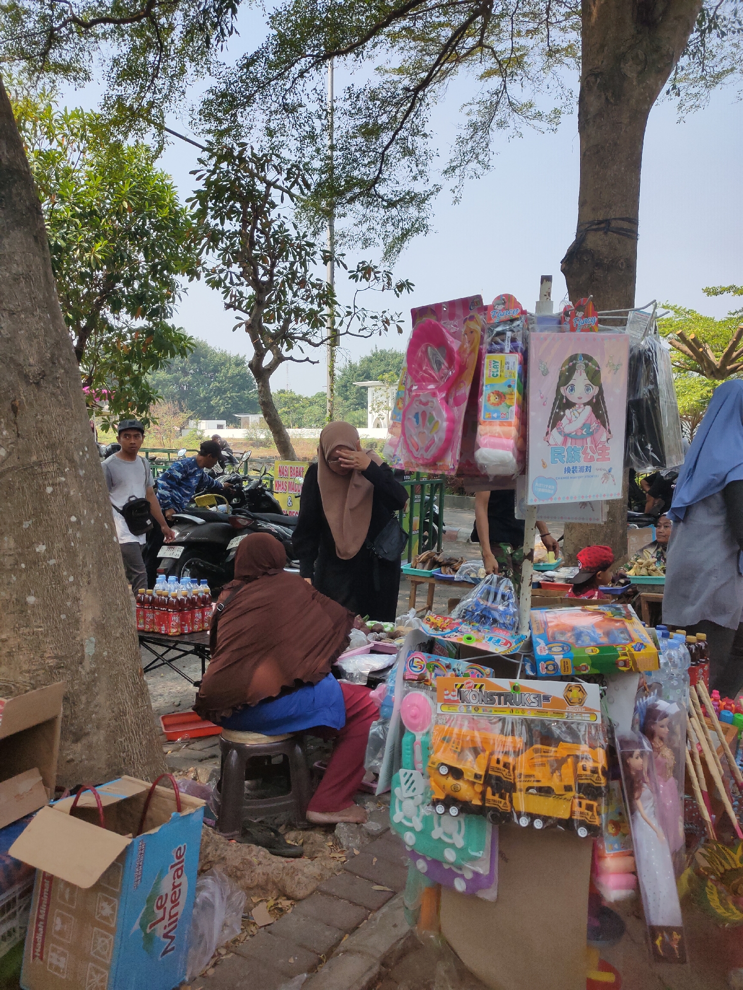 The image captures a street market scene in Ngasem, Indonesia, under a canopy of trees. A vendor, wearing a brown hijab, sits on a stool behind a display of bottled drinks and other goods. In front of her, a young child with a red cap is partially visible. Adjacent to the drinks, various toys are arranged on display, including construction vehicles and dolls. To the left, a man wearing a grey t-shirt and a black cap stands near a motorcycle. In the background, more people and motorcycles are visible, along with a building and dense greenery. The ground is paved with bricks.