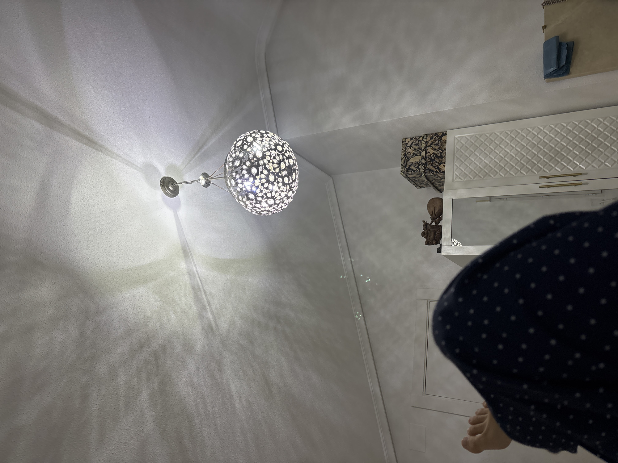 The image shows a bedroom interior with an upward-angle view towards a sloped ceiling. A spherical chandelier with intricate cutouts hangs from the ceiling, casting dappled light patterns on the textured white surface. In the foreground, a person's legs are visible, covered by dark blue pajamas with white polka dots, suggesting they are lying down. To the right, a white wardrobe with gold handles stands against the wall, topped with two decorative boxes and a small wooden figurine. The room appears to be in an apartment or house in Orihuela, Spain.