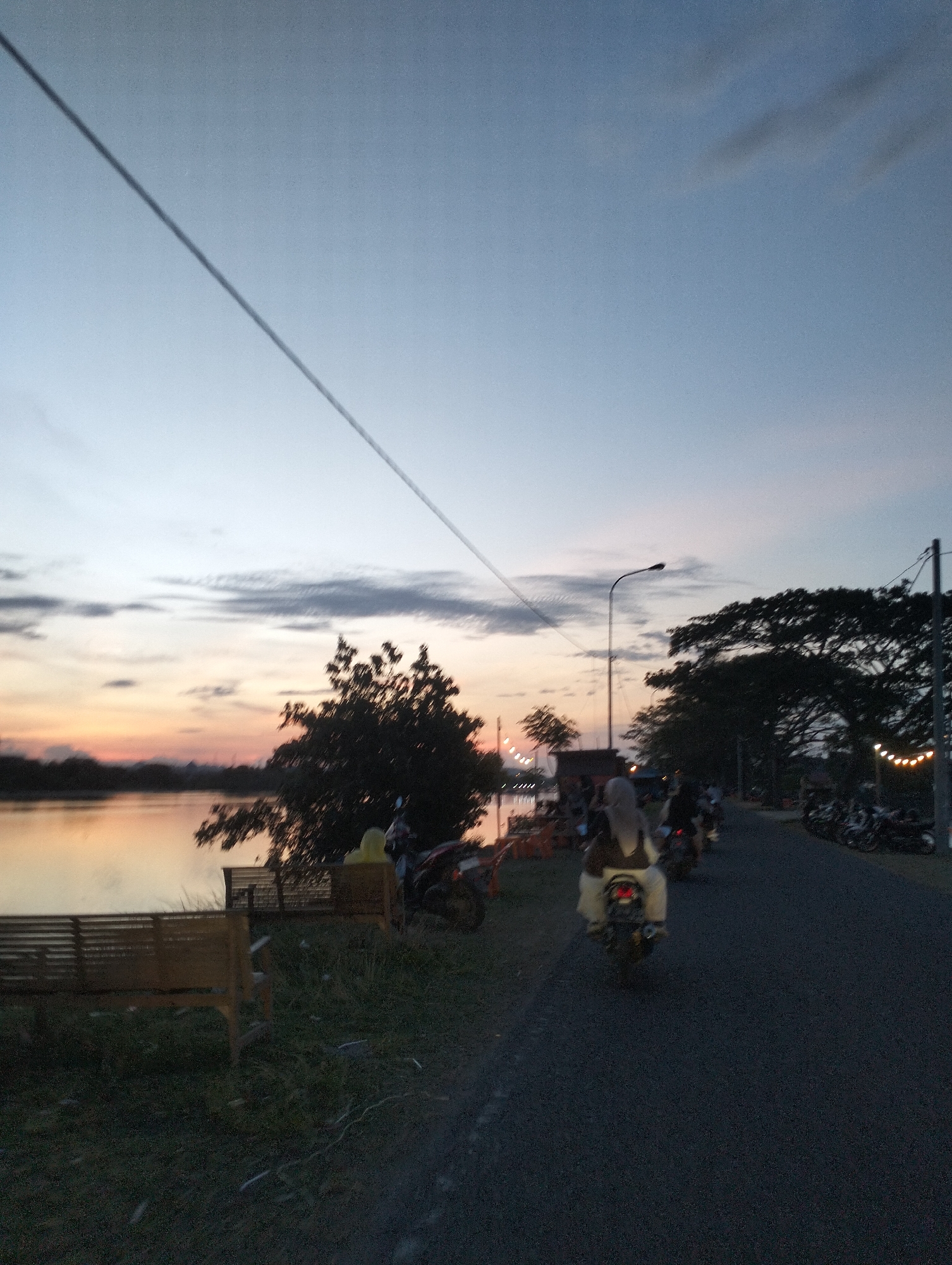 The image depicts a lakeside view at dusk in Lhokseumawe, Indonesia. The sky is a mix of light blue and orange hues reflecting the sunset. A body of water, possibly a lake, is visible on the left side of the frame. There are some trees and vegetation on the bank of the lake, with a wooden bench visible.
A road runs along the right side of the image. Several motorbikes, with riders, are on the road. A street lamp is visible along with trees that have string lights. There are also parked motorbikes further down the road. The lighting conditions suggest it is either late evening or early morning.