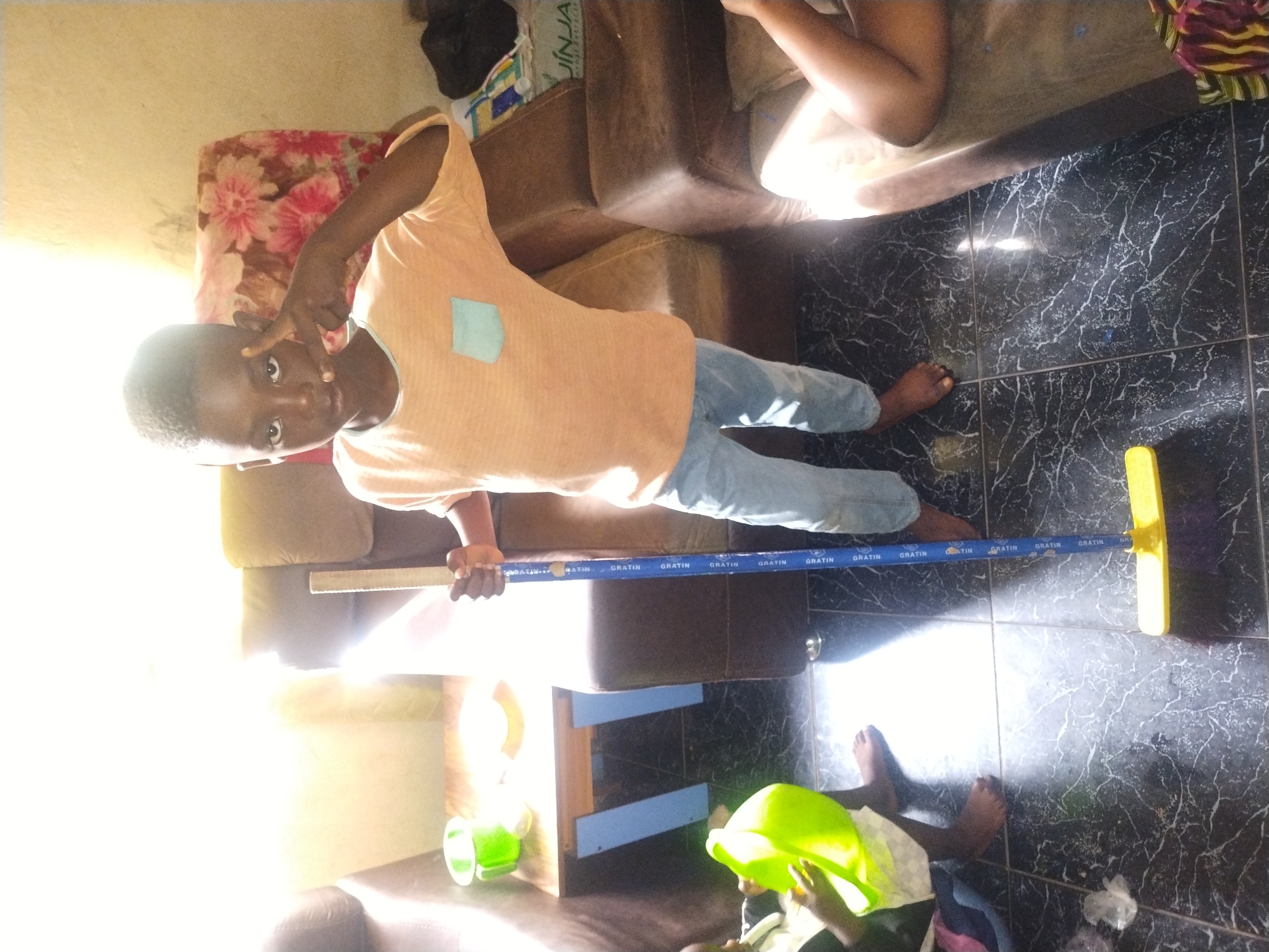 An eye-level, medium shot captures a young boy standing indoors, holding a yellow-bristled broom with a blue handle. The boy, with dark skin and hair, wears a light orange t-shirt and blue jeans. He faces the camera with his left hand raised in a peace sign. The floor is tiled in black with white veins. To the left of the boy, another child, mostly out of frame, sits on the floor holding a bright green bucket. In the background, a brown leather couch is visible, with a floral-patterned cloth draped over its back. A small table with a green cup and a white plate sits behind the couch. Natural light streams in from a window on the left, creating a bright glare.