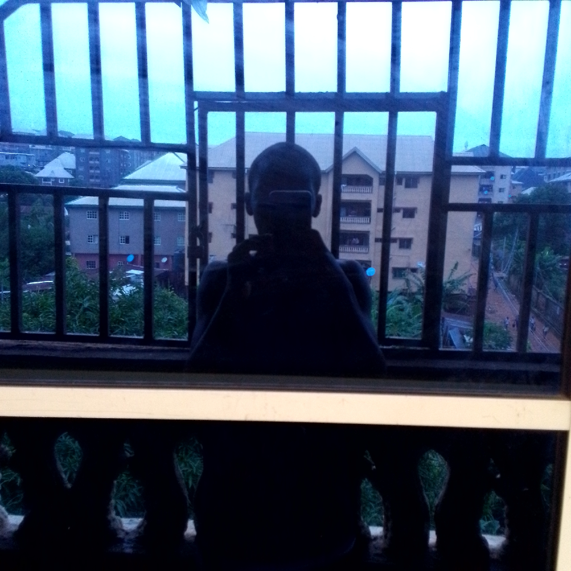 The image is a self-portrait taken indoors, reflected in a window with a blue tint. The photographer is holding a phone to capture the reflection. Through the window, a cityscape with multiple buildings and greenery is visible. The buildings appear to be residential, with balconies and visible windows. The sky is overcast and light blue. In the foreground, below the window, is a dark, reflective surface showing the lower part of the photographer's body and the railing of what seems to be a balcony. The overall lighting suggests it is daytime, likely in the early morning or late afternoon due to the soft light and overcast conditions.