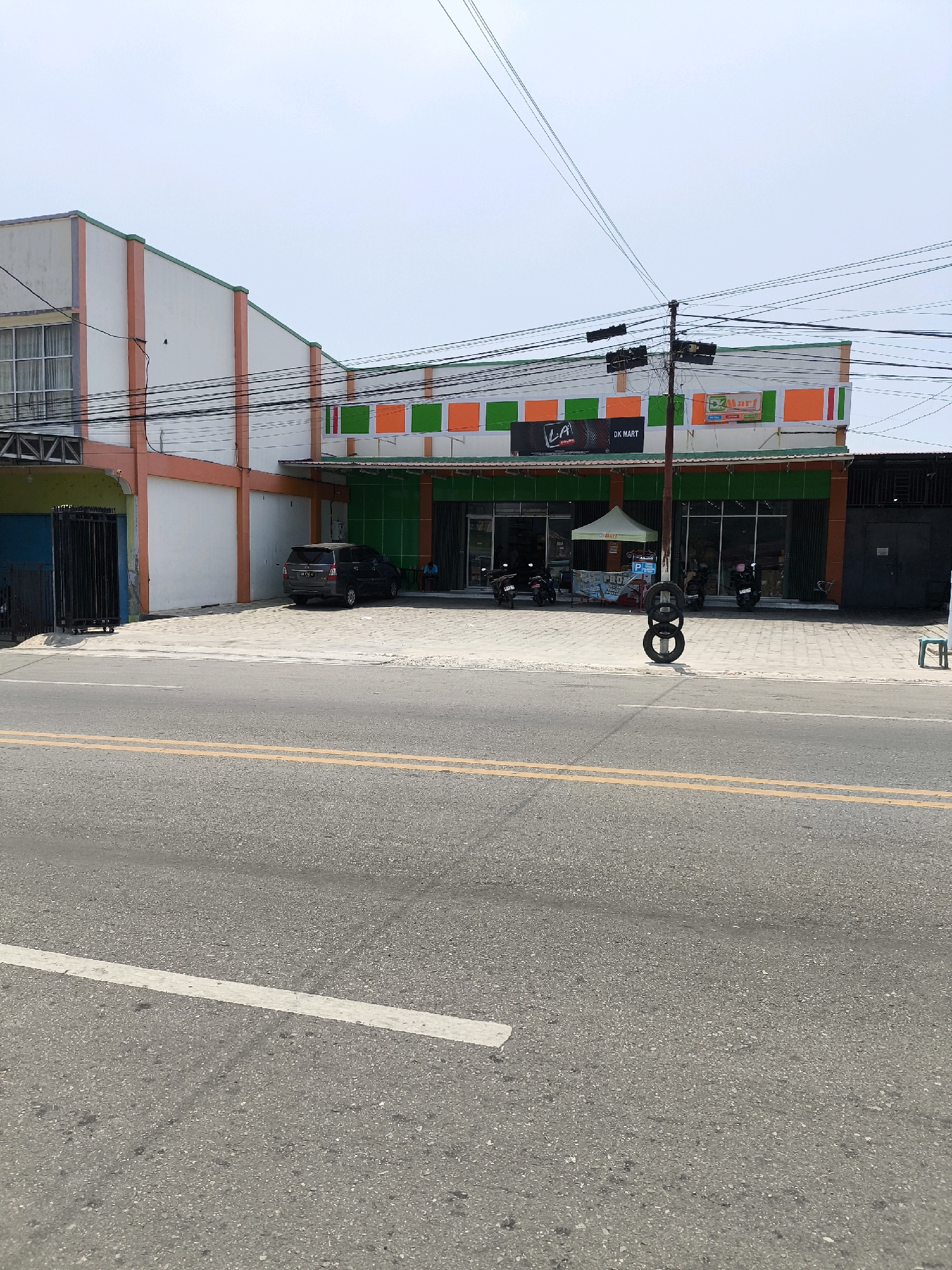 The image shows a street-level view of a DK Mart convenience store in Kwandang, Indonesia. The store has a white facade with orange and green accents. The entrance is marked by a green awning and glass doors. There are several motorcycles parked outside, and a dark-colored minivan is parked in front of the building. A person is sitting on a stool outside the store. In the foreground, there is a street with yellow lane markings. A stack of tires is visible on the sidewalk in front of the store. The sky is overcast.
