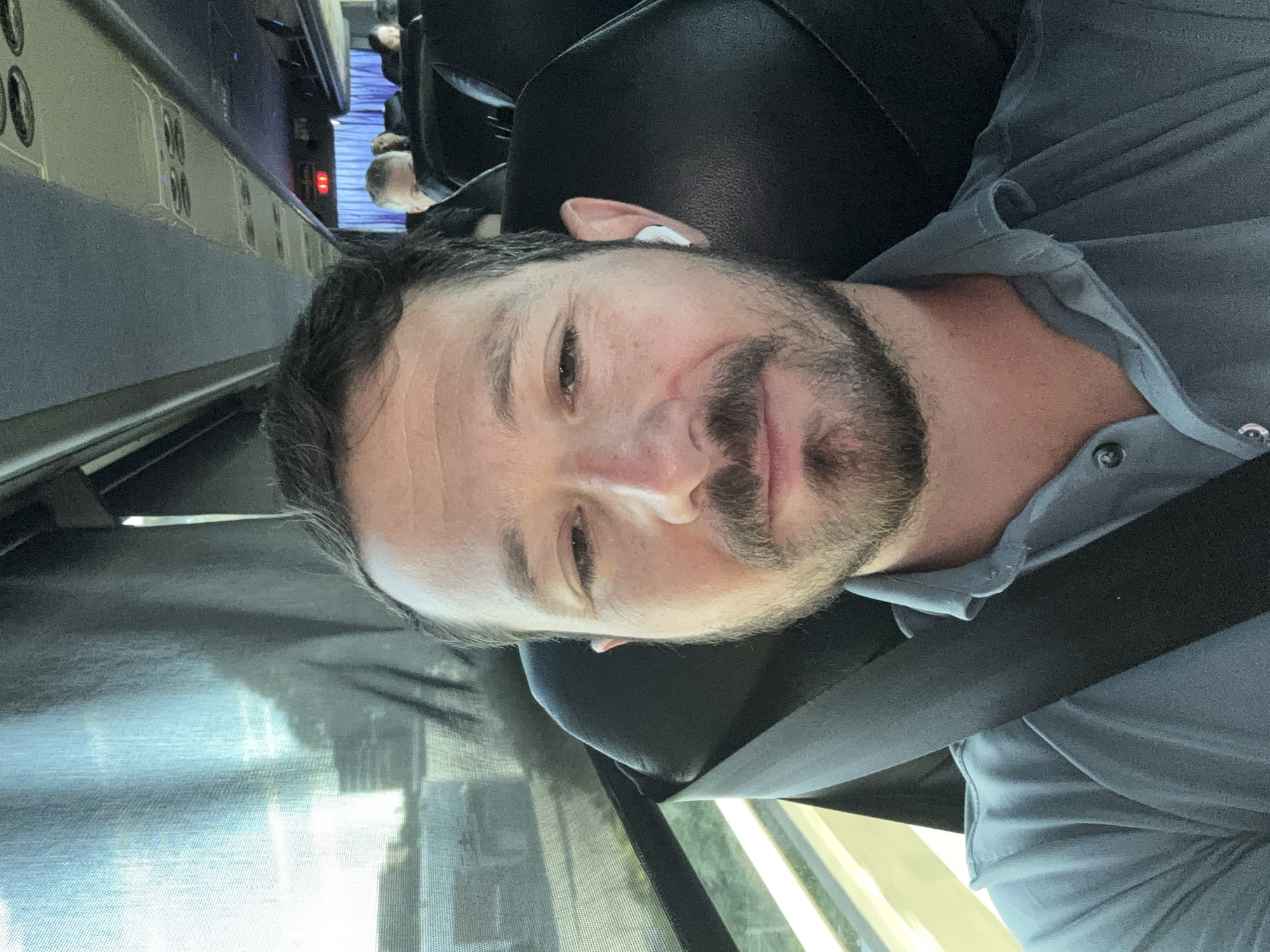 A male passenger is depicted in a selfie taken inside a bus or shuttle. The man, appearing to be in his 30s or 40s, has short brown hair and a full beard, and is wearing a light blue collared shirt. A black seatbelt is diagonally secured across his chest and left shoulder. A white wireless earbud is present in his right ear. He is seated in a black upholstered seat. Through the bus's left-side window, obscured by a dark mesh screen, a blurred view of a highway or road with green vegetation and a distant light-colored structure is visible, indicative of a daytime setting. In the background, other passengers are partially visible in additional black seats along the aisle, with blue curtains and red indicator lights observed on the ceiling towards the rear of the vehicle. The specific location within San Mateo, United States of America, cannot be determined from the provided image.
