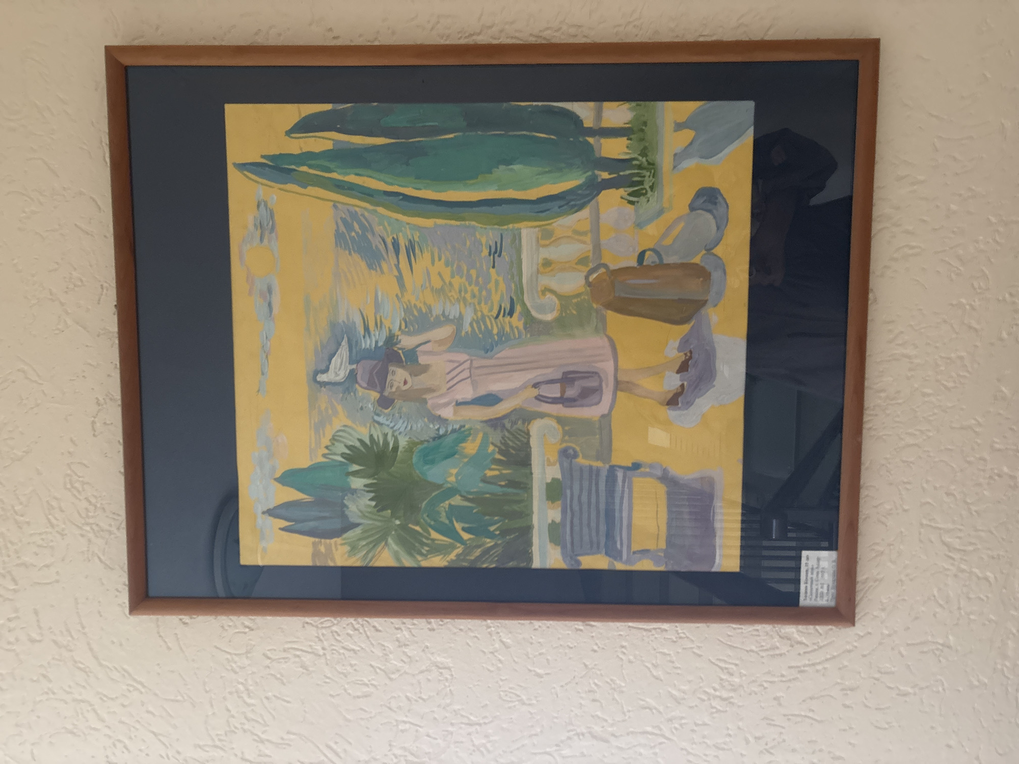 A framed painting hangs on a textured, off-white wall. The painting depicts a scene in a style reminiscent of Impressionism or Post-Impressionism. The main subject is a woman in a dress and hat, lying down in a garden setting. There are stylized trees, foliage, and architectural elements such as a balustrade. The color palette includes yellow, green, blue, and lavender tones. There is a label on the lower right corner of the painting. Based on the label text, the painting depicts a scene from the city of Sochi, Russia.