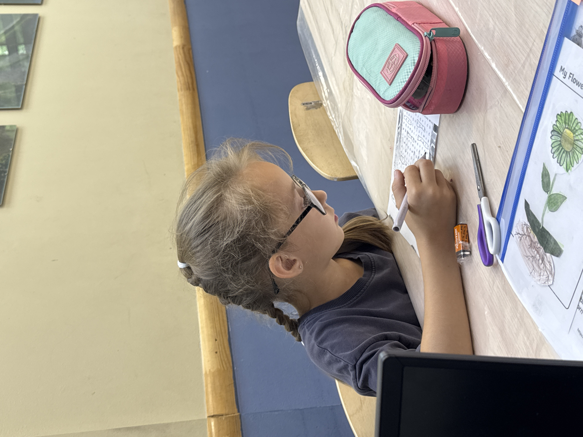 A young girl with blonde hair in a braid, wearing glasses and a dark grey t-shirt, is sitting at a wooden table and drawing. She is focused on a piece of paper in front of her. On the table, there is a pink and teal pencil case, a pair of purple and white scissors, a battery, and a laptop. In the background, there is a wall painted in two tones, a wooden shelf, and framed pictures on the wall. The setting appears to be an indoor educational or recreational space. There is no indication of the city of Perm, Russia, or any specific geographical location.