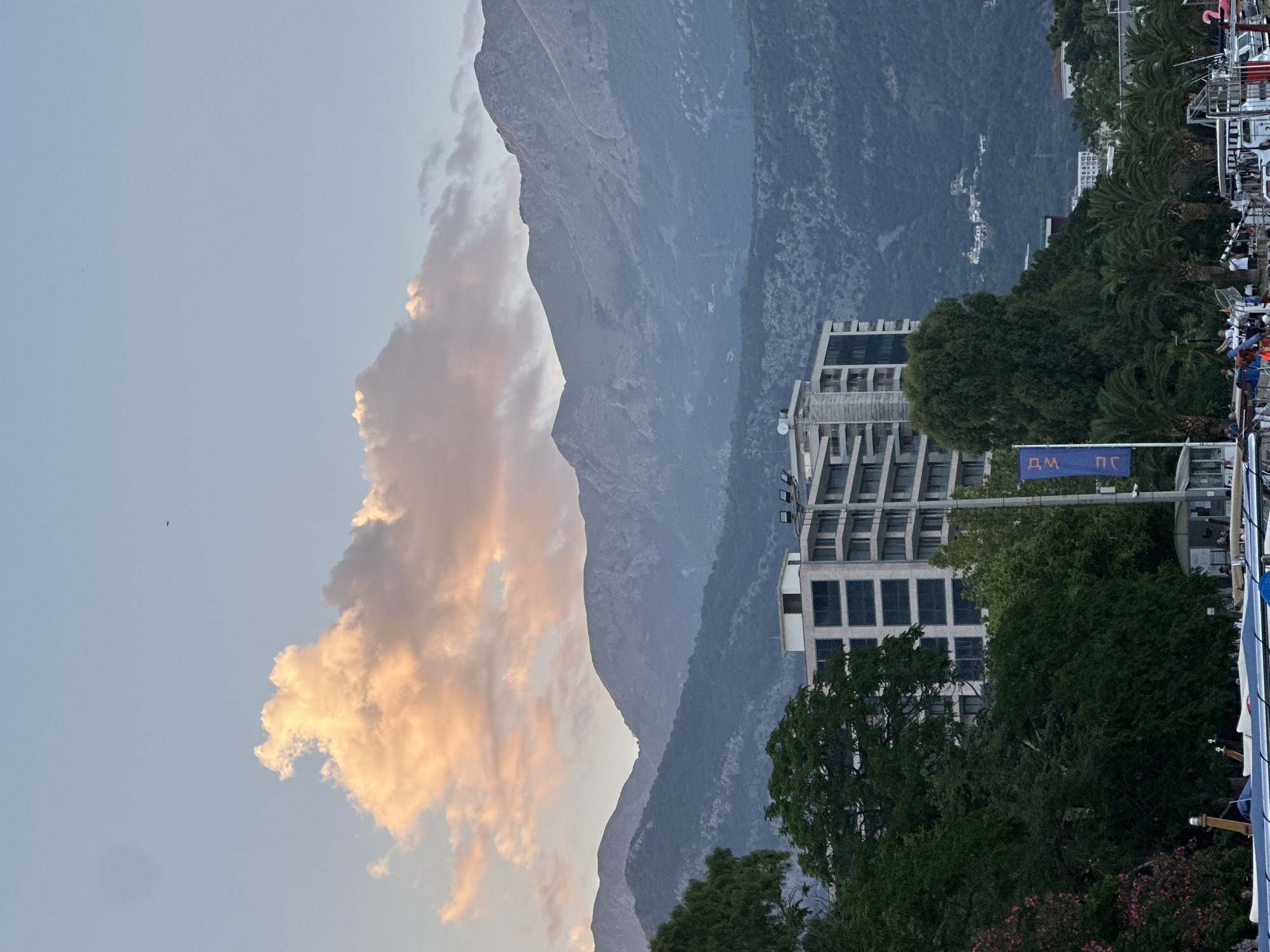 The image captures a scenic view of Budva, Montenegro, dominated by the majestic mountains that form the backdrop to the coastal city. In the foreground, a modern, multi-story building with a white facade and numerous windows stands amidst lush green trees. The building appears to be a hotel or apartment complex, suggesting an urban or resort setting. Palm trees are visible, adding to the Mediterranean atmosphere. A blue flag with white lettering is planted near the building, possibly indicating a local establishment or event. In the distance, the rugged, verdant slopes of the mountains rise, their peaks softened by the evening light. The sky above is a pale blue, transitioning into a soft pink and orange hue near a large, fluffy cloud, indicating either sunrise or sunset. Several boats are docked in the lower part of the image, hinting at a harbor or waterfront area. A few people are visible on the docks, engaged in various activities. The overall scene is serene and picturesque, blending natural beauty with urban development.