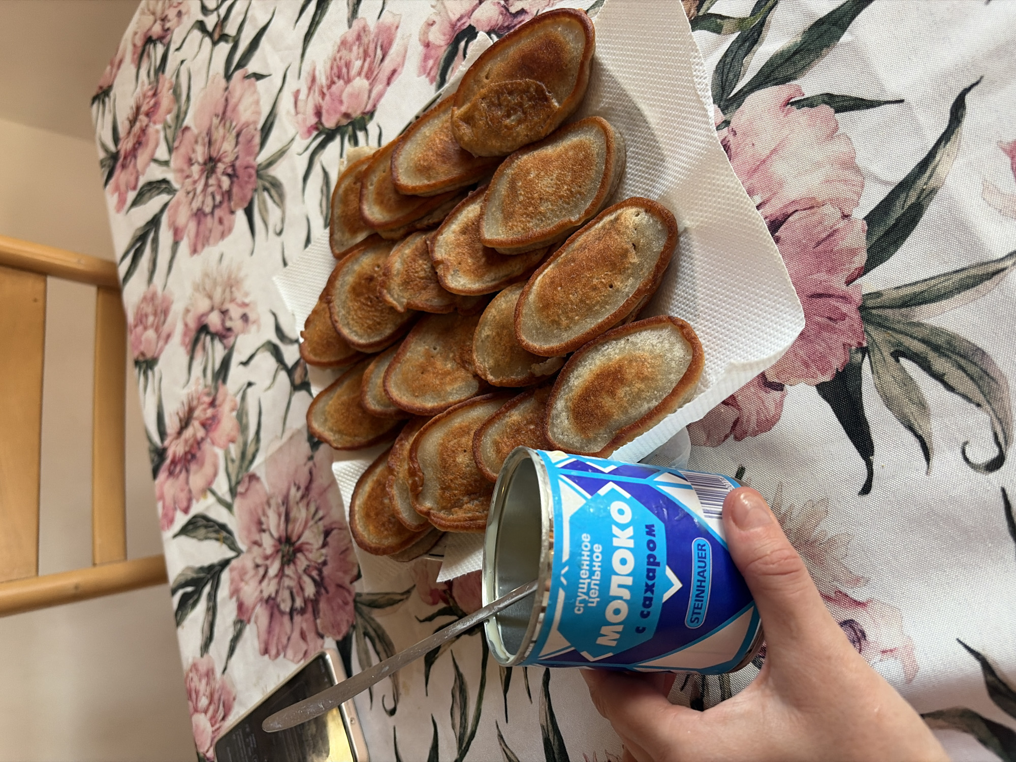 A hand holds a can of condensed milk with a spoon inside, positioned to the left of a plate of small, oval-shaped pancakes. The pancakes are golden brown and arranged in rows on a white paper napkin. The background features a floral-patterned tablecloth with large pink peonies and green leaves. A wooden chair is partially visible at the top of the frame. The scene appears to be indoors, on a table. The location is not specified in the image.
