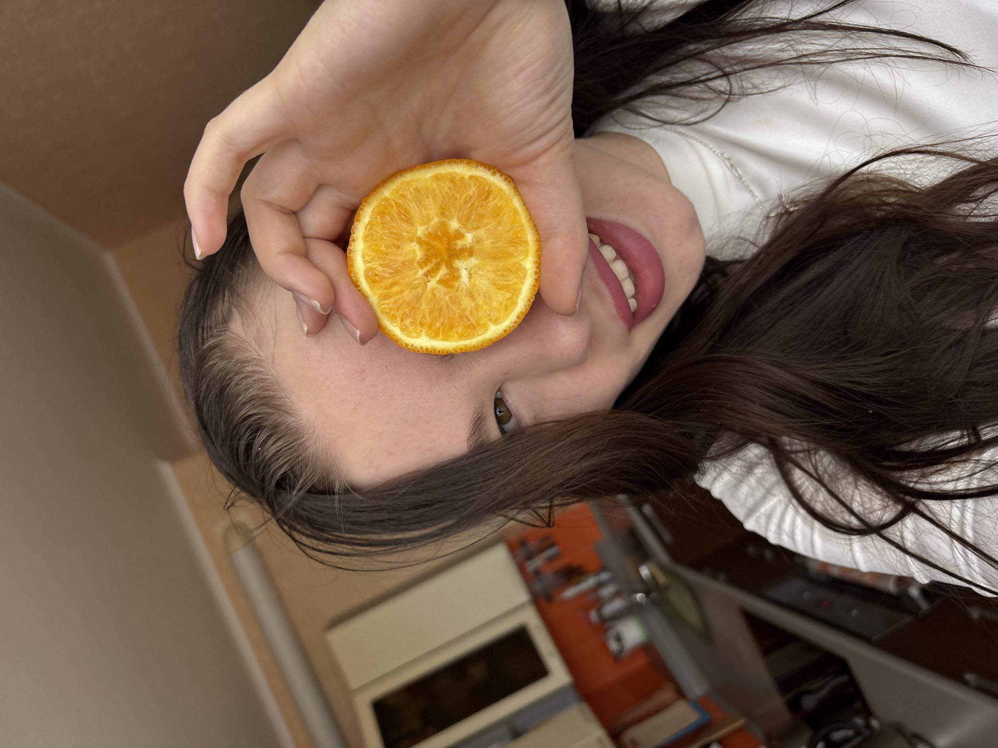 A woman is holding a slice of orange in front of her eye. She has dark hair and fair skin. She is wearing a white shirt. She is partially visible. The background shows a kitchen setting with cabinets and countertops. The photograph was taken indoors. The city and country are not discernible from the image.