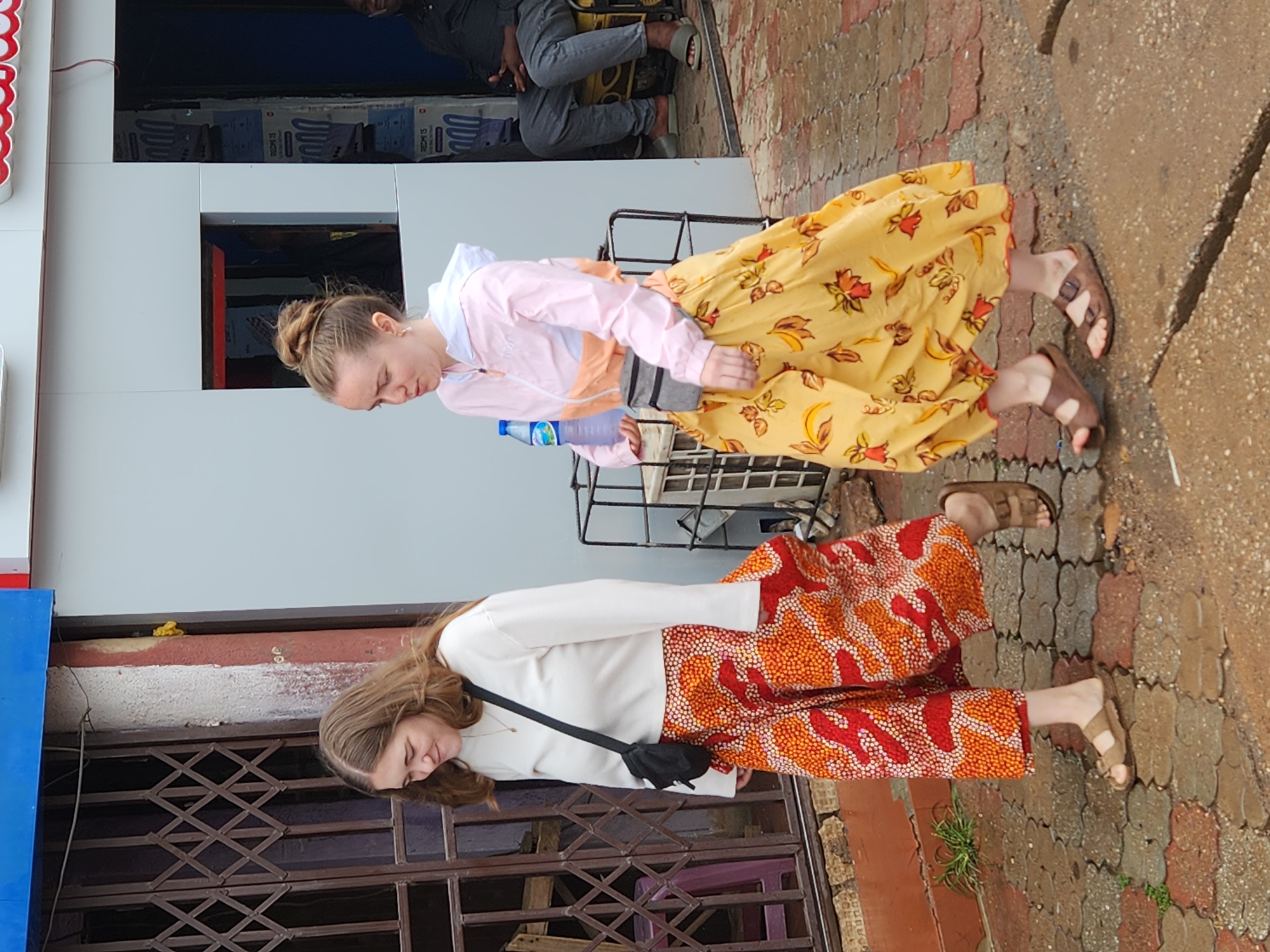 Two women are standing on a brick walkway in front of a building. The woman on the left wears a long-sleeved white shirt and patterned orange and red pants, with a black bag slung over her shoulder. The woman on the right wears a pink jacket, a yellow dress with a floral pattern, and sandals. She carries a water bottle. A metal grate is partially visible in the background on the left, and a white structure with a window is behind the other woman. A person is sitting on a yellow object in the background.