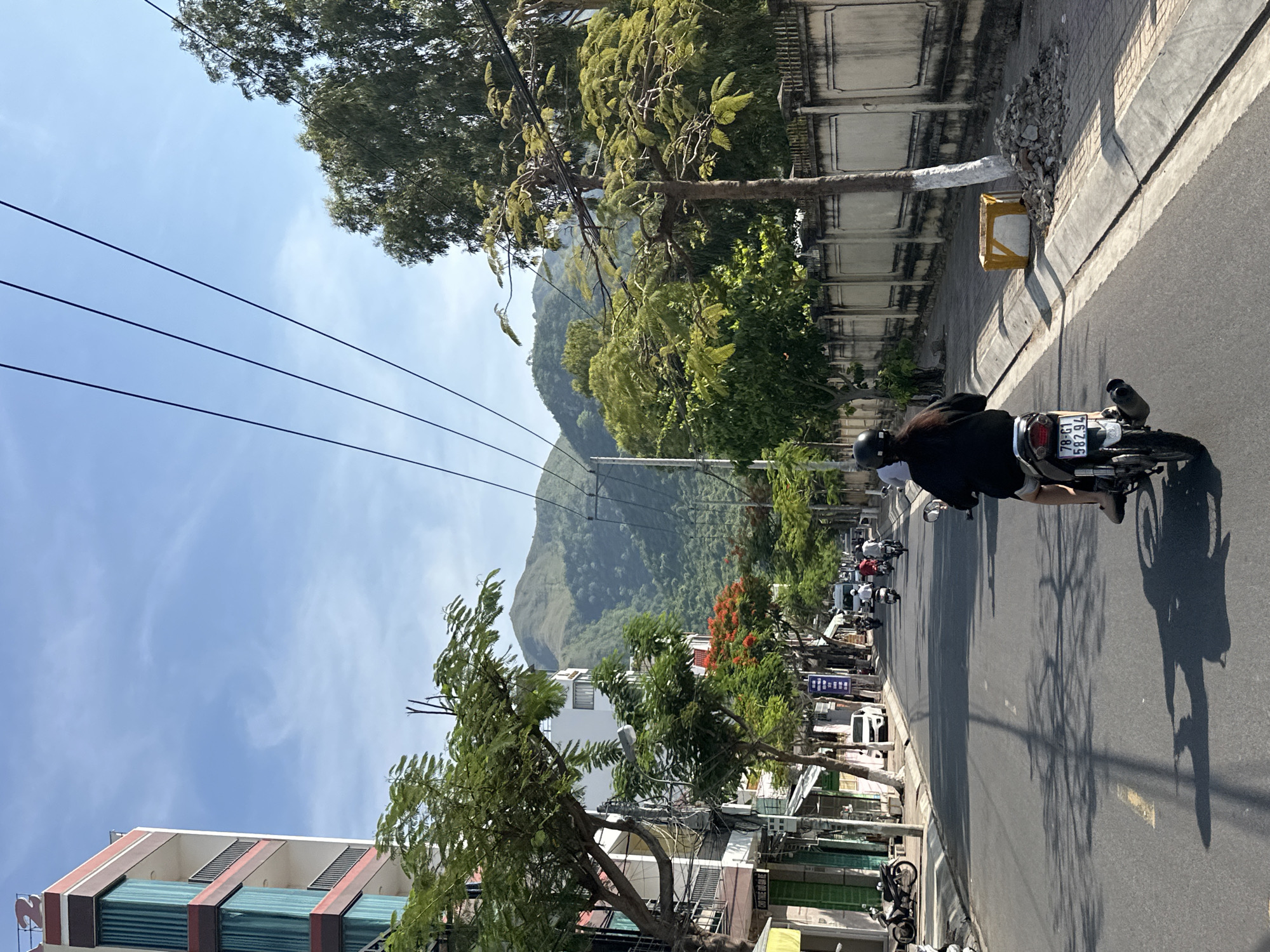 The image depicts a street scene in Bac Nha Trang Ward, Vietnam. A person wearing a black jacket and helmet rides a motorcycle down a paved road. The motorcycle has a license plate reading "78-G1 582.94". The road is lined with buildings on the left and a wall with trees on the right. In the background, lush green mountains are visible under a clear blue sky with some wispy clouds. Power lines stretch across the frame above the street. Several other motorcycles are also present on the road. The overall impression is a sunny day in a moderately populated urban area with natural mountainous terrain in the distance.
