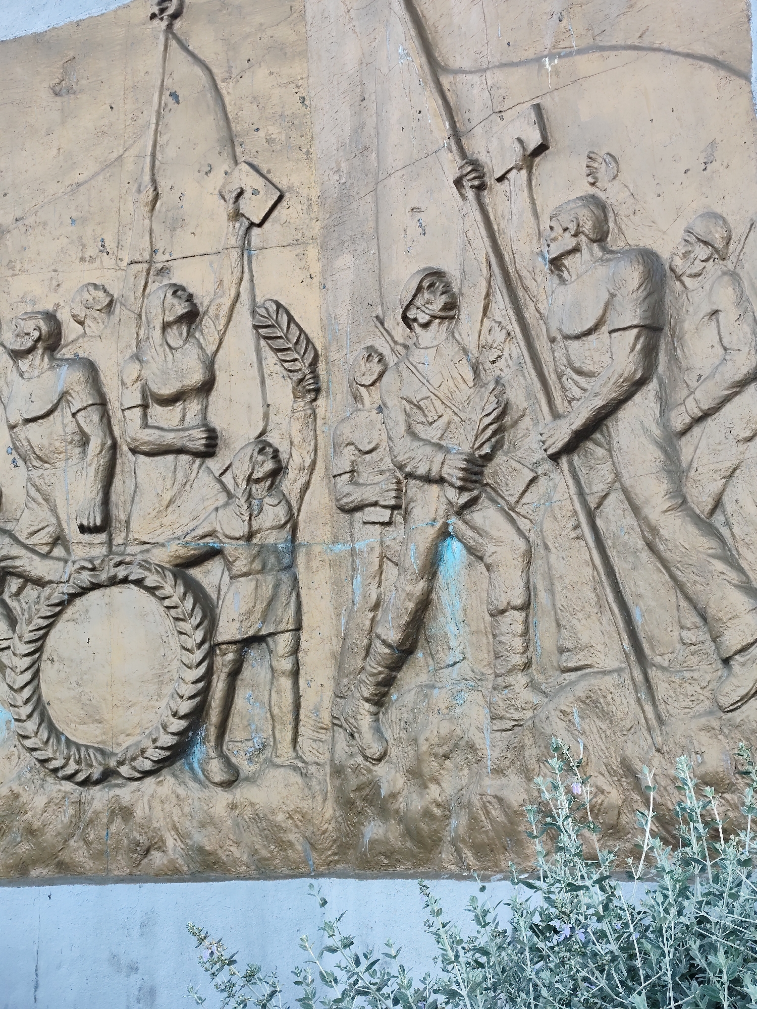 The image shows a bronze relief sculpture mounted on a wall in an urban environment, presumably outdoors. The sculpture depicts a group of figures, with a style reminiscent of socialist realism, engaged in a collective action or procession. They are portrayed as determined and forward-moving. Details of the figures include soldiers in helmets, workers, and individuals holding symbols like a flag or a laurel wreath, possibly representing struggle and victory. The surface of the relief shows signs of weathering and age. The bottom part of the image features plant life, with some flowering plants growing against the wall. The exact location is specified as Unknown, Unknown.