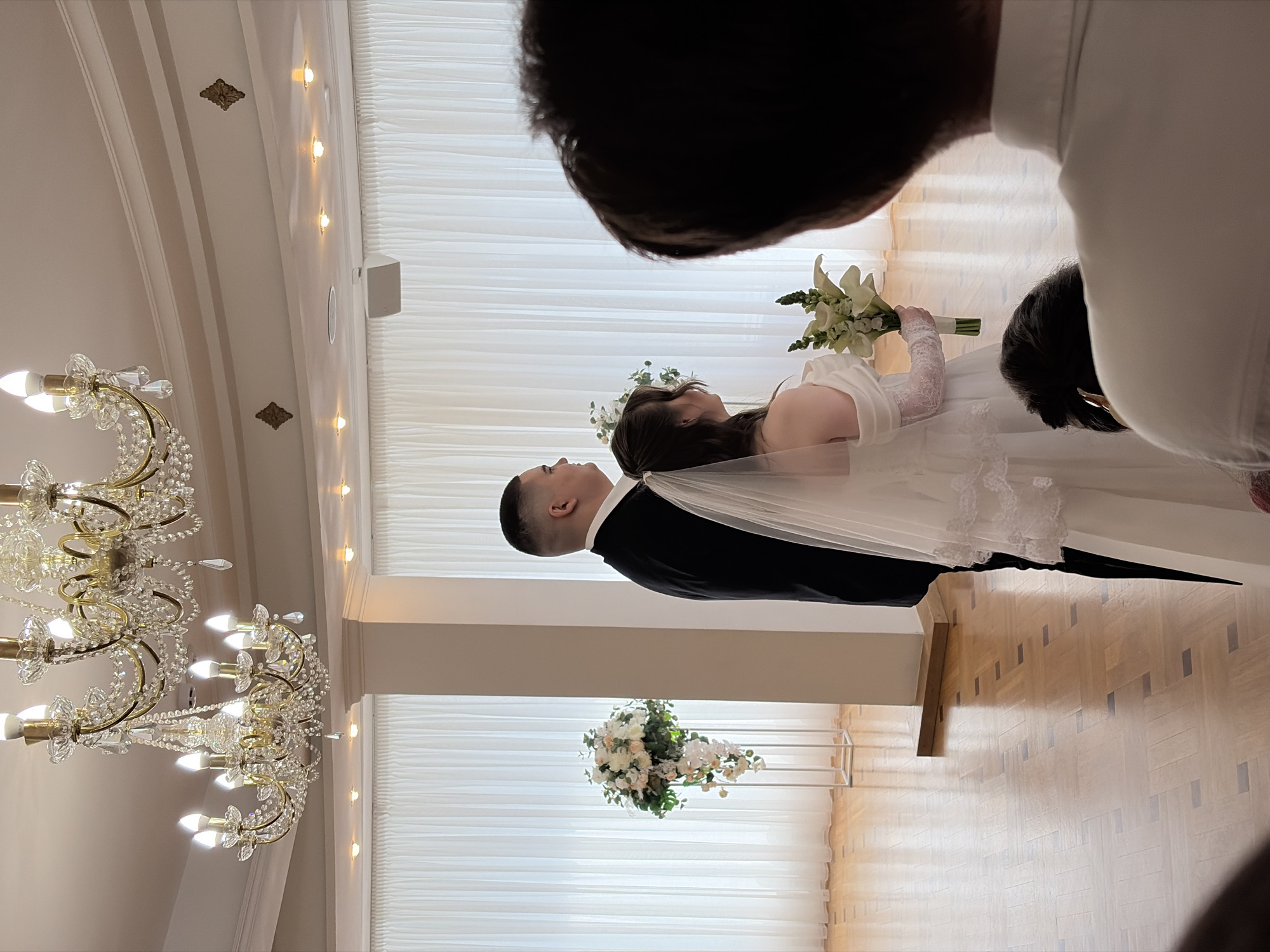 A wedding ceremony is in progress indoors. A groom wearing a black suit and a bride in a white gown and veil are facing each other. The bride is holding a bouquet of white lilies. The setting includes white draped curtains, floral arrangements, a chandelier, and a wood-patterned floor. Two individuals are standing in the foreground, partially obstructing the view of the couple. The location is possibly in Yekaterinburg, Russia, if that information is verified independently.