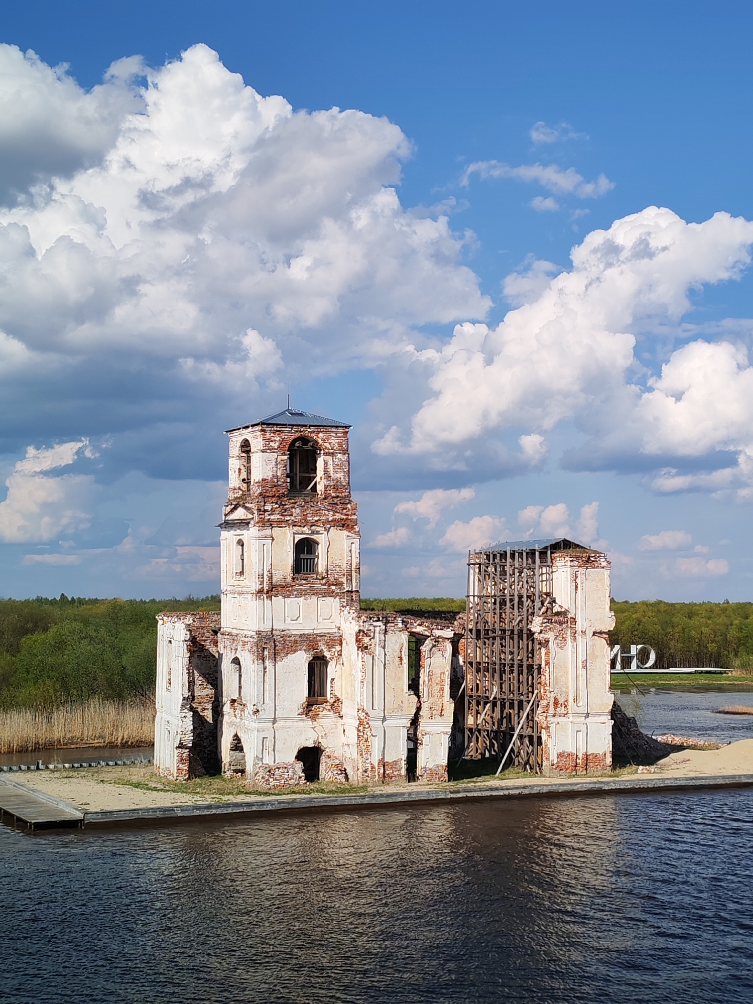 The image depicts the ruined Church of the Nativity in Krokhino, Russia. The dilapidated structure, built of white and red brick, stands in the middle of a body of water. The church's roof is partially intact, with scaffolding supporting one section. The surrounding environment includes a shoreline with green trees and reeds. The sky is bright blue with numerous white clouds.