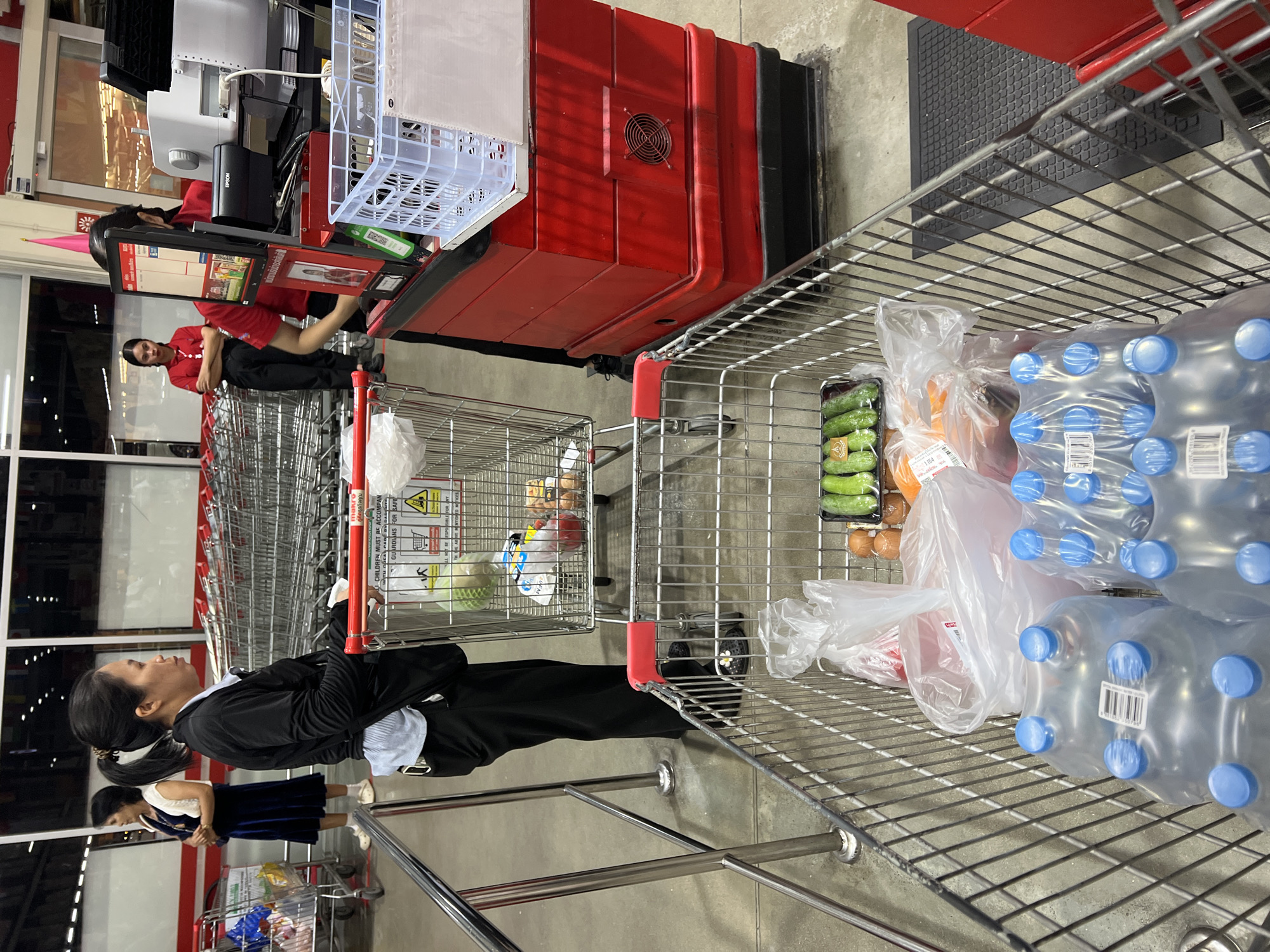 In Baan Bo Phut, Thailand, a woman with her hair pulled back in a ponytail is seen from the side, wearing a black jacket over a light blue and white striped shirt, and black pants. She is standing next to a shopping cart filled with groceries. The cart contains several large bottles of water with blue caps, plastic bags of produce including cucumbers, and some other items. Behind her, a second shopping cart is partially visible, loaded with various goods. In the background, two women in red shirts appear to be working at a checkout counter. One is looking at a screen, and the other has her arms crossed. Numerous shopping carts are neatly stacked in the distance, suggesting a retail environment, likely a supermarket or hypermarket. The overall scene depicts a typical retail shopping experience.