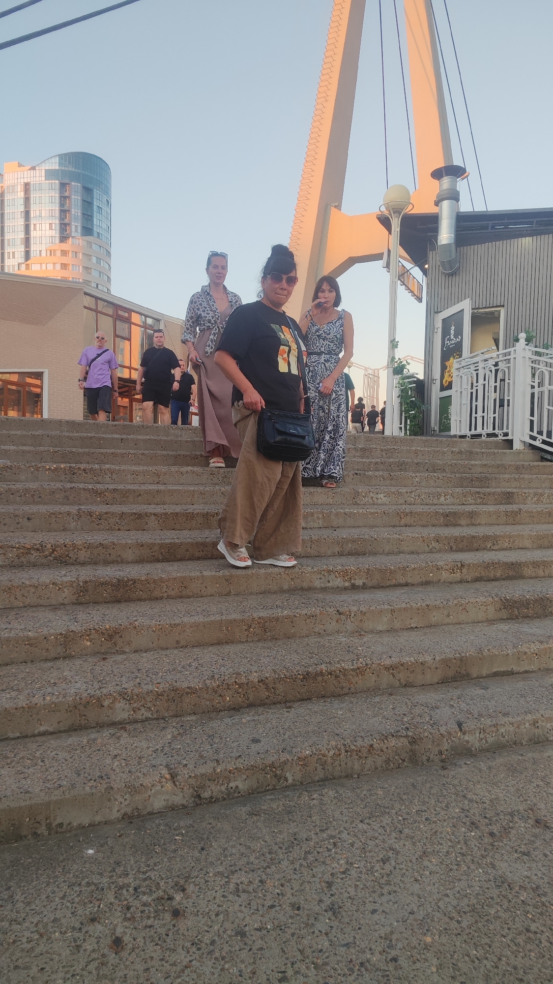 The image shows a group of people on a concrete staircase. The scene appears to be outdoors, with a clear sky and what looks like a building with large windows in the background. There is also a large, light-colored structure resembling a bridge or a large support beam in the upper right portion of the image. The people are dressed in casual clothing. The overall setting seems to be an urban environment. The question of location in Krasnodar, Russia, is not supported by any visual cues in the image.