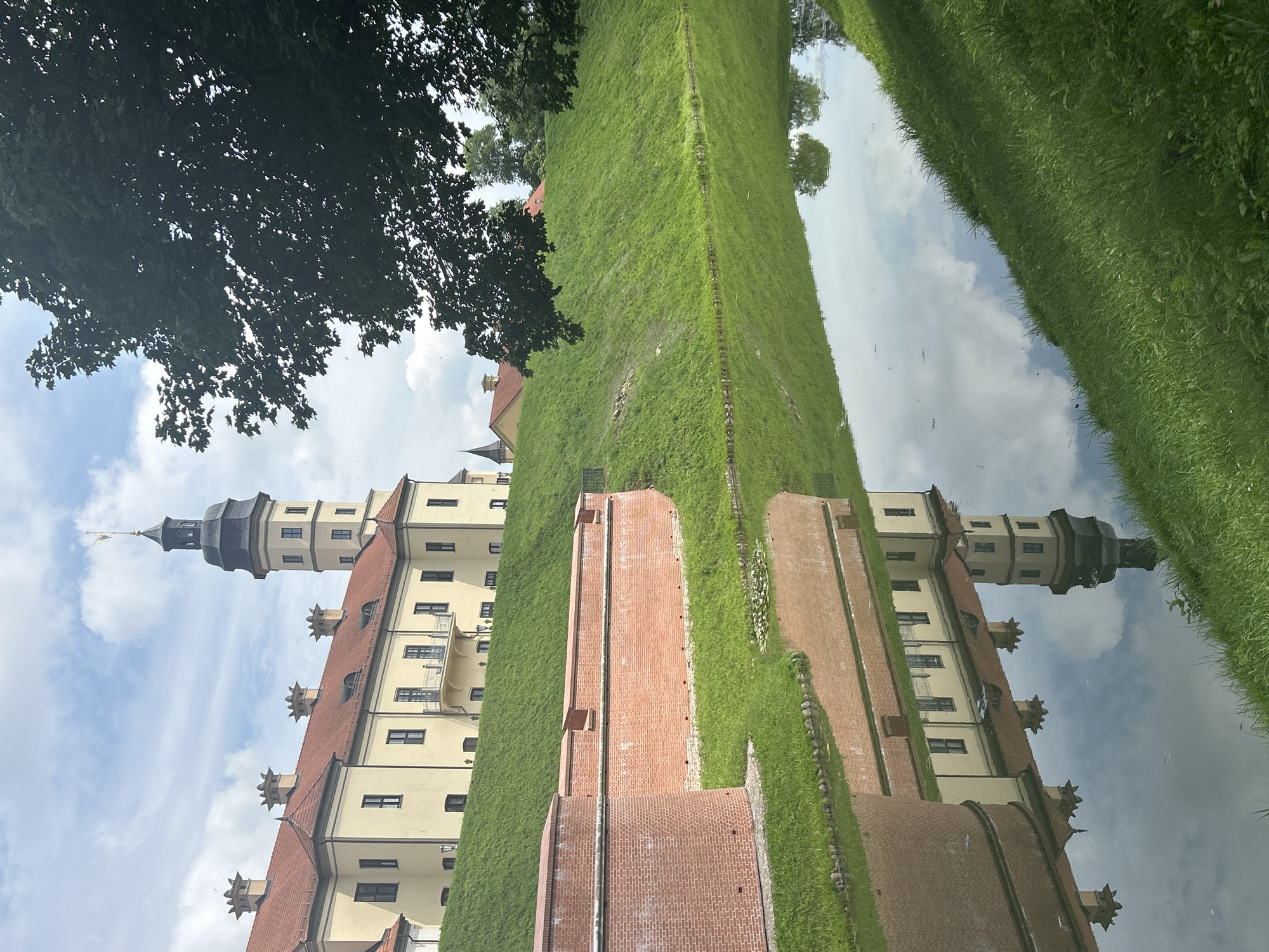 The image shows a picturesque view of Nesvizh Castle in Nesvizh, Belarus. A large, light yellow building with a red-tiled roof and multiple towers stands on a grassy hill. In the foreground, a moat reflects the castle and the sky. A brick wall with battlements borders the moat and the lower part of the hill. Lush green grass covers the slopes and the area around the moat. The sky is partly cloudy with a mix of blue sky and white clouds. A large tree with dark green leaves partially obstructs the view on the right side of the image. The scene is serene and suggests a historical and well-maintained tourist attraction. There are no people visible in the image.