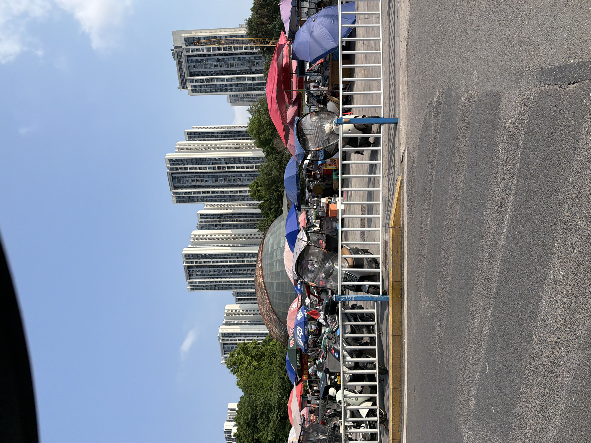 A street scene in Chongqing High-tech Zone, China, shows a row of market stalls lining the sidewalk, each covered with colorful umbrellas for shade. The stalls appear to sell various items, though specific products are difficult to discern. A white metal fence separates the market area from the road. Behind the stalls, a cluster of tall, modern apartment buildings rises against a clear blue sky. The road appears to be asphalt.