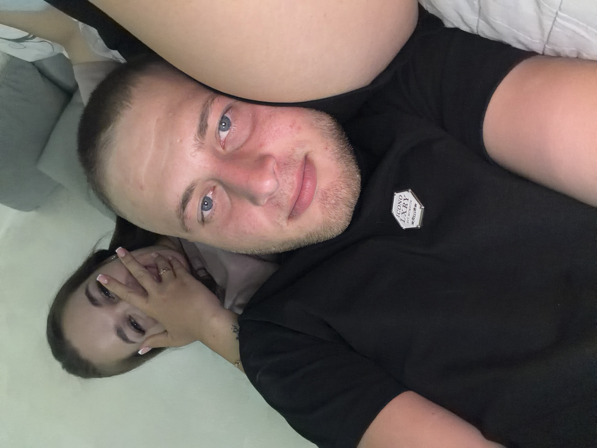 Two individuals, a male and a female, are lying down. The male, positioned in the foreground, has blue eyes and a light beard. He is wearing a black t-shirt with a hexagonal logo. The female, positioned behind the male, is partially obscured by her hand covering her mouth. She has dark hair and is wearing a light-colored t-shirt. A ring is visible on her left hand. They appear to be in a domestic setting, possibly a bedroom, based on the presence of pillows and a wall in the background. The image is taken from a close-up perspective, with the subjects filling most of the frame. There is no discernible information to place this image specifically in the city of Strășeni, Moldova.