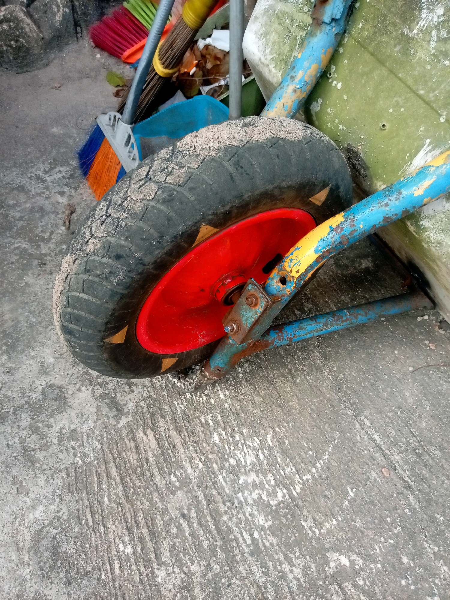 A close-up, high-angle shot captures a worn, red and blue wheelbarrow's wheel resting on a rough, gray concrete surface. The tire is dirty, with sand clinging to its tread. A red rim is visible beneath the tire. The wheelbarrow's blue and yellow painted frame is visible, showing signs of rust and wear. To the left of the wheelbarrow, a blue dustpan and parts of several brooms with orange, red, and green bristles are present, suggesting a cleaning area or storage of cleaning supplies. The background shows a portion of a weathered, greenish object, possibly another piece of equipment or a wall. The scene appears to be outdoors, in what could be a service area or a yard within Batam, Indonesia.