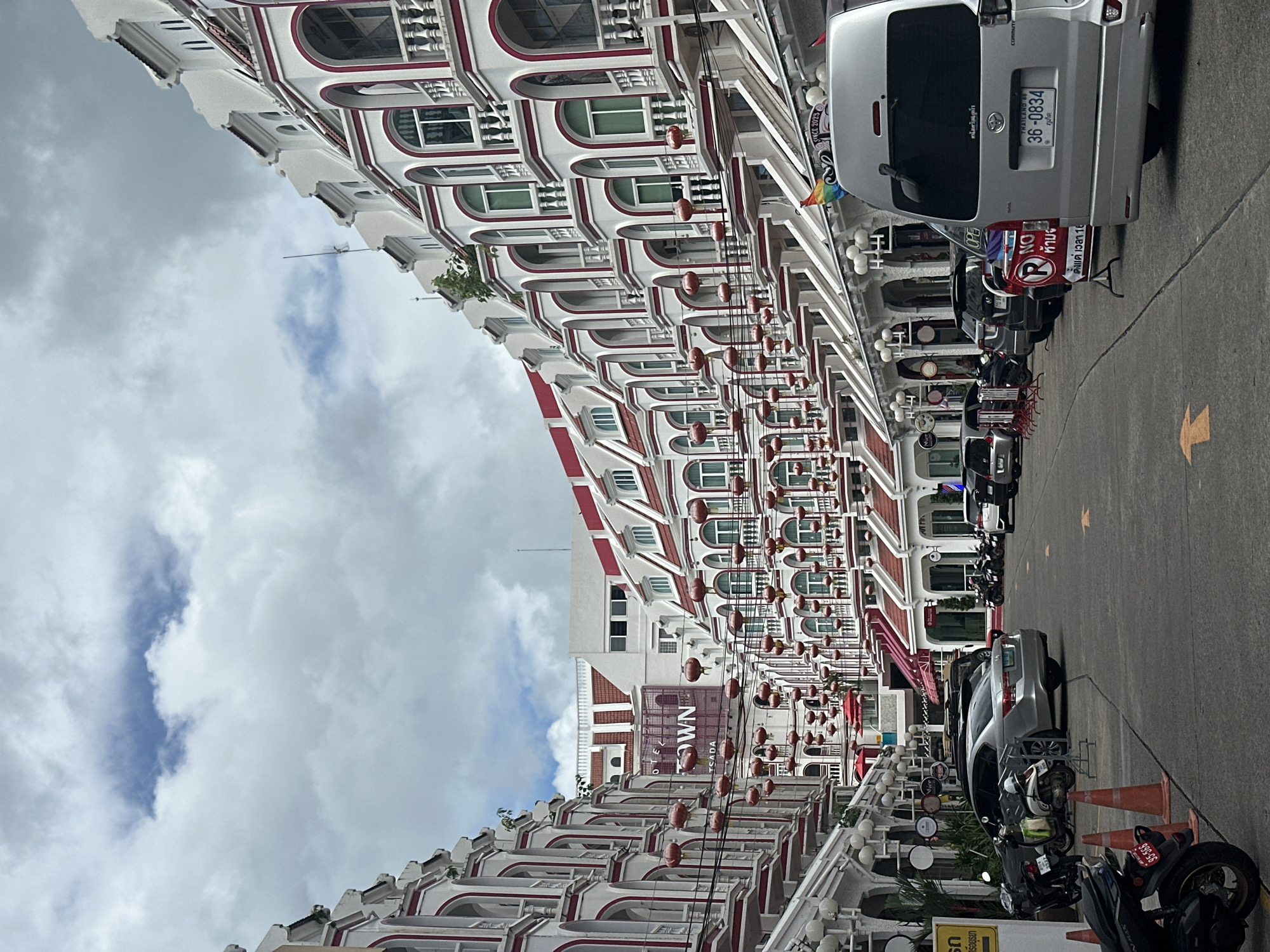 This image shows a street scene in Phuket City Municipality, Thailand. The perspective is tilted, giving a dynamic feel to the composition. The left side of the image is dominated by a building with a distinctive architectural style, featuring rows of arched windows with red trim and white decorative elements. Red lanterns are strung along the front of the building, suggesting a festive or cultural ambiance. On the right side, several vehicles are parked, including a silver van with a Thai license plate reading "36-0834", a silver car, and several motorcycles. One motorcycle has a license plate that reads "95-566". Signs are visible, including one with "NO" and a red circle with a slash, indicating a prohibition. Another sign reads "TOWN". The sky is cloudy, suggesting overcast weather. The overall scene depicts a street in a town, likely in an urban or commercial area, with a mix of traditional architecture and modern vehicles. The time of day appears to be daytime, given the lighting.
