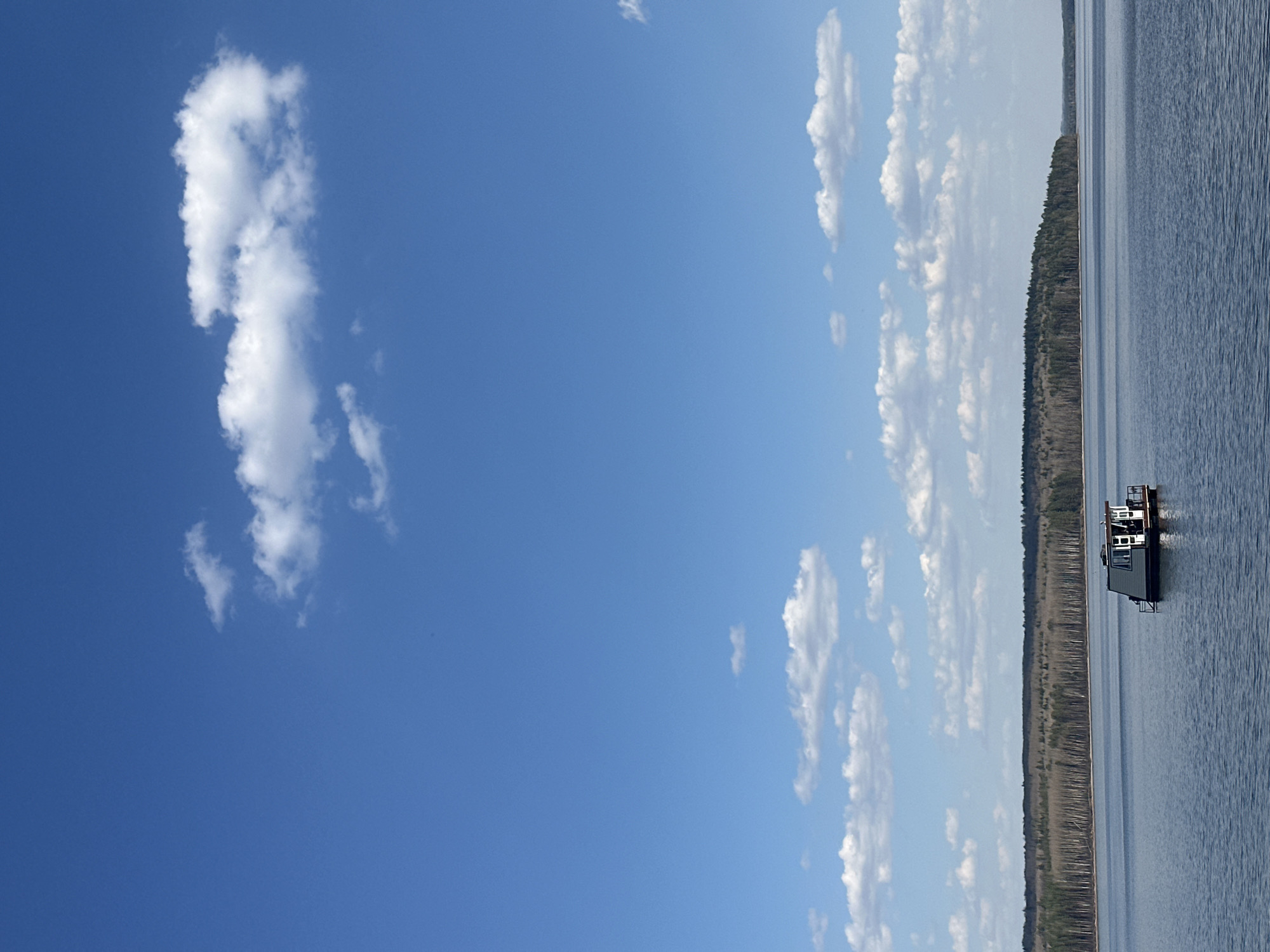 The image shows a body of water, likely a lake or reservoir, under a blue sky with scattered clouds. A shoreline with trees runs along the right side of the water. There is a floating structure, possibly a boathouse or a small building on a platform, visible on the water near the shoreline. The location is likely in Chaikovsky, Russia, given the context provided. The scene appears to be a calm, sunny day in a natural or semi-natural setting.