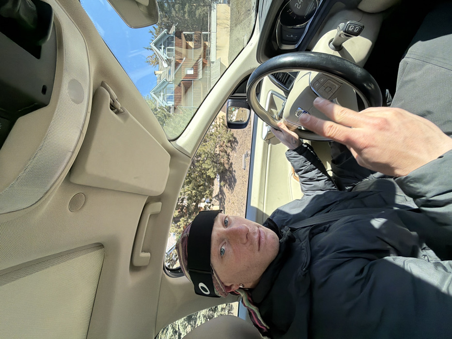 A male individual is seated in the driver's seat of a Volvo vehicle on a sunny day in La Salle-les-Alpes, France. He has light skin, blue eyes, and a neutral expression, looking directly at the viewer. He wears a black Oakley headband over a multicolored patterned headwrap, a silver hoop earring in his left ear, and a dark, possibly black or grey, puffer jacket. His right hand is positioned on the steering wheel, while his left hand is raised in front of the steering wheel, displaying an extended index and middle finger gesture. A small tattoo is visible on his left wrist. The vehicle's interior features a beige dashboard and ceiling, a sunroof, and a sun visor. The instrument cluster shows a numerical reading of "24" and "D" for the gear. Through the passenger-side window and windshield, a multi-story chalet-style house with light brown wooden siding and distinctive blue-grey gabled roofs is visible, partially obscured by pine trees. The background also reveals a clear blue sky and some dry ground patches.
