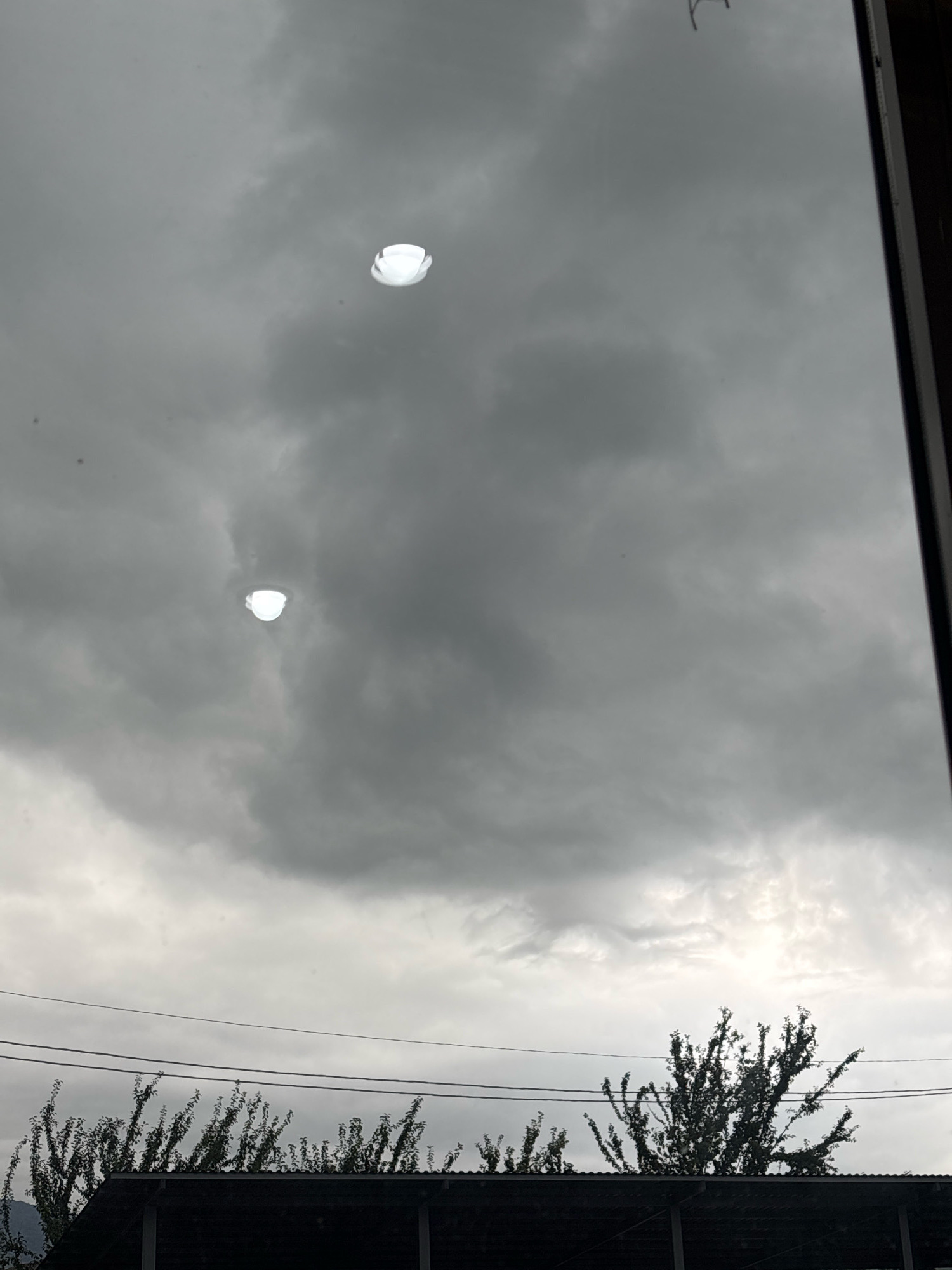 This is a photograph taken in Ijevan, Armenia, looking upwards through a window. The foreground of the image is dominated by a dark roof structure and silhouetted trees, with power lines stretching across the sky. The sky itself is overcast and grey, with dramatic cloud formations. Two bright, white, disc-shaped objects are visible in the sky, appearing as reflections on the windowpane, not as actual objects in the sky. These reflections are illuminated and stand out against the dark clouds. The overall impression is a somewhat somber atmosphere due to the cloudy sky and the dark elements in the foreground.