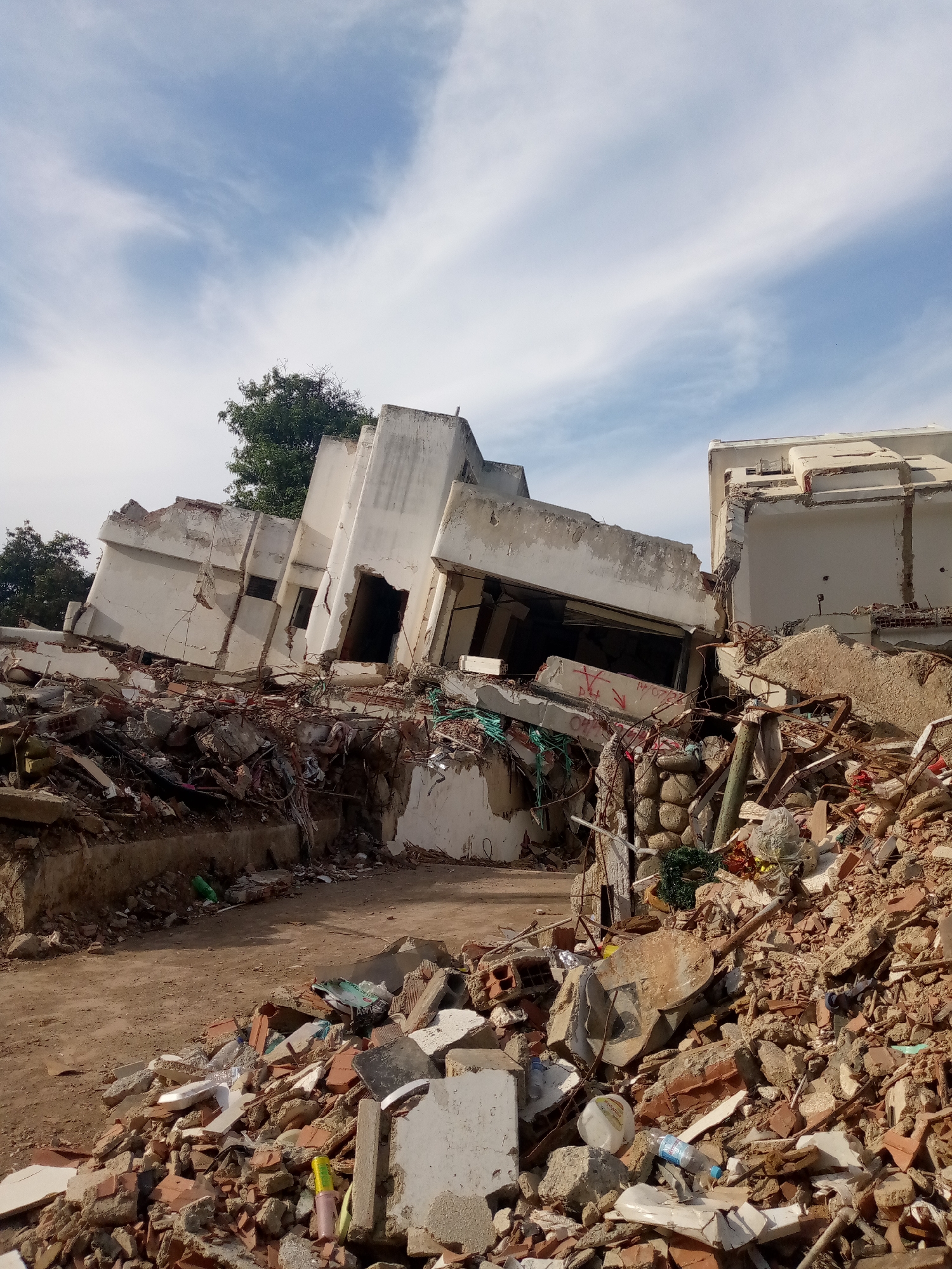 The image depicts the aftermath of a significant disaster, likely a landslide or earthquake, in the city of Caraballeda, Venezuela. A multi-story building has collapsed, with its structure severely damaged and tilted at a precarious angle. Large sections of the building are in ruins, with debris scattered everywhere. Piles of concrete, bricks, rebar, and other building materials are strewn across the ground, creating an uneven and hazardous landscape.
The front of the building shows gaping holes where walls and windows once were. Exposed rebar protrudes from the broken concrete, and tangled wires are visible amidst the rubble. The upper floors have crumbled, with sections of the roof and walls lying in heaps. In the foreground, the ground is covered with smaller debris, including shattered bricks and pieces of concrete. Some plastic bottles and other discarded items are mixed in with the rubble, suggesting the area may have been inhabited or used for storage.
A single green tree stands tall behind the damaged structure, its leaves providing a stark contrast to the destruction. The sky above is a pale blue with scattered white clouds. The overall scene is one of devastation and chaos, indicative of a catastrophic event that has significantly impacted the urban environment. The presence of such widespread destruction suggests a major natural disaster.